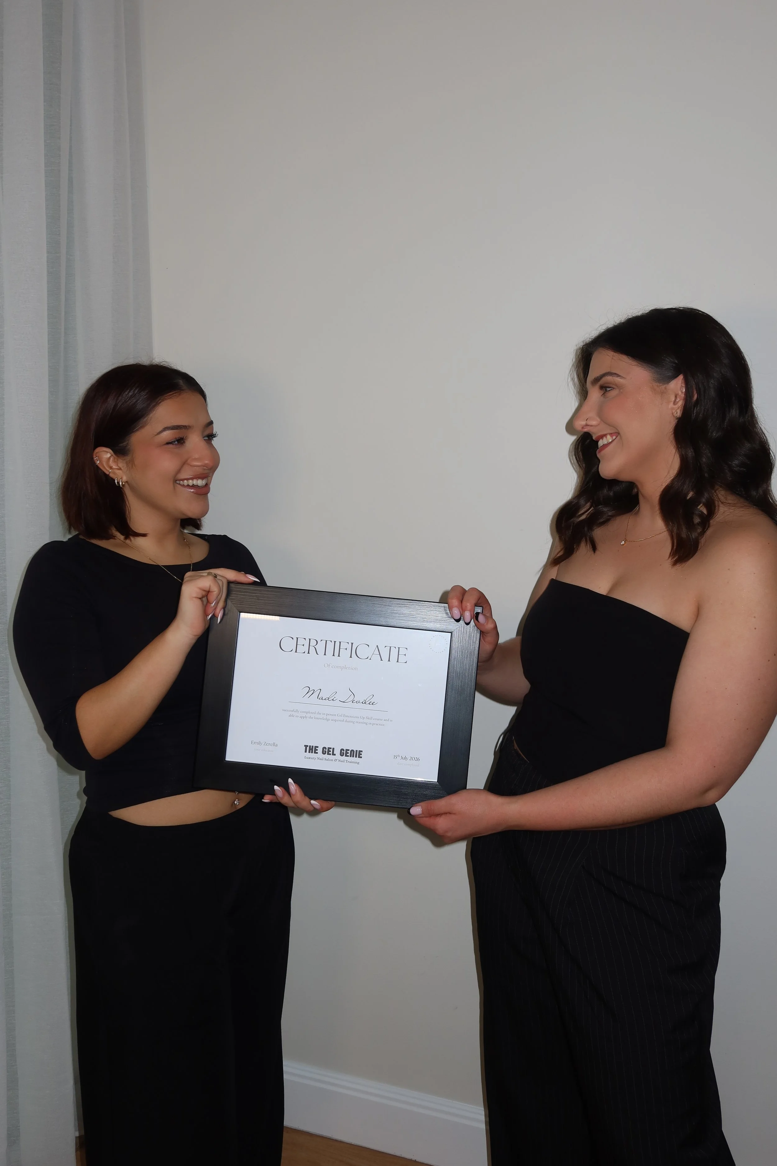 Two women standing indoors, smiling and exchanging a framed certificate, with a white wall and window with sheer curtains in the background.