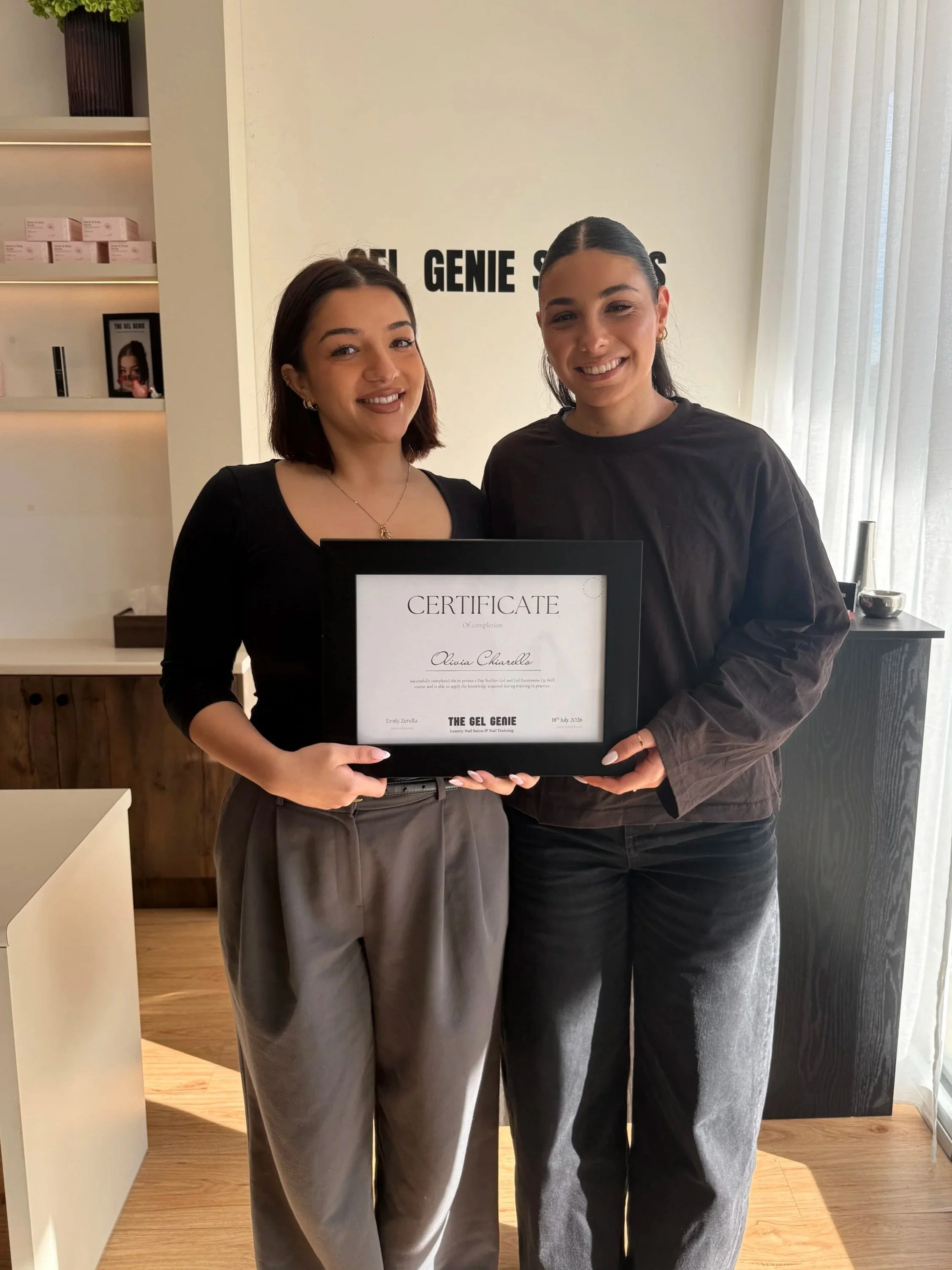 Two women smiling and holding a certificate in a modern room with light-colored walls and curtains. Shelf with pink boxes and framed picture are in the background.