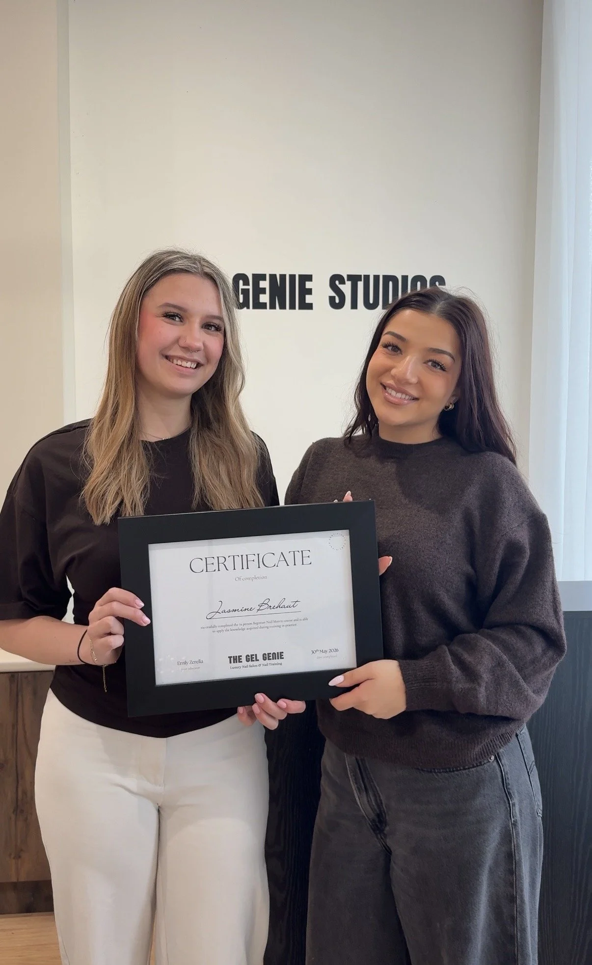 Two women standing indoors, holding a framed certificate together, smiling at the camera. Behind them is a wall with the text 'GENIE STUDIOS'.
