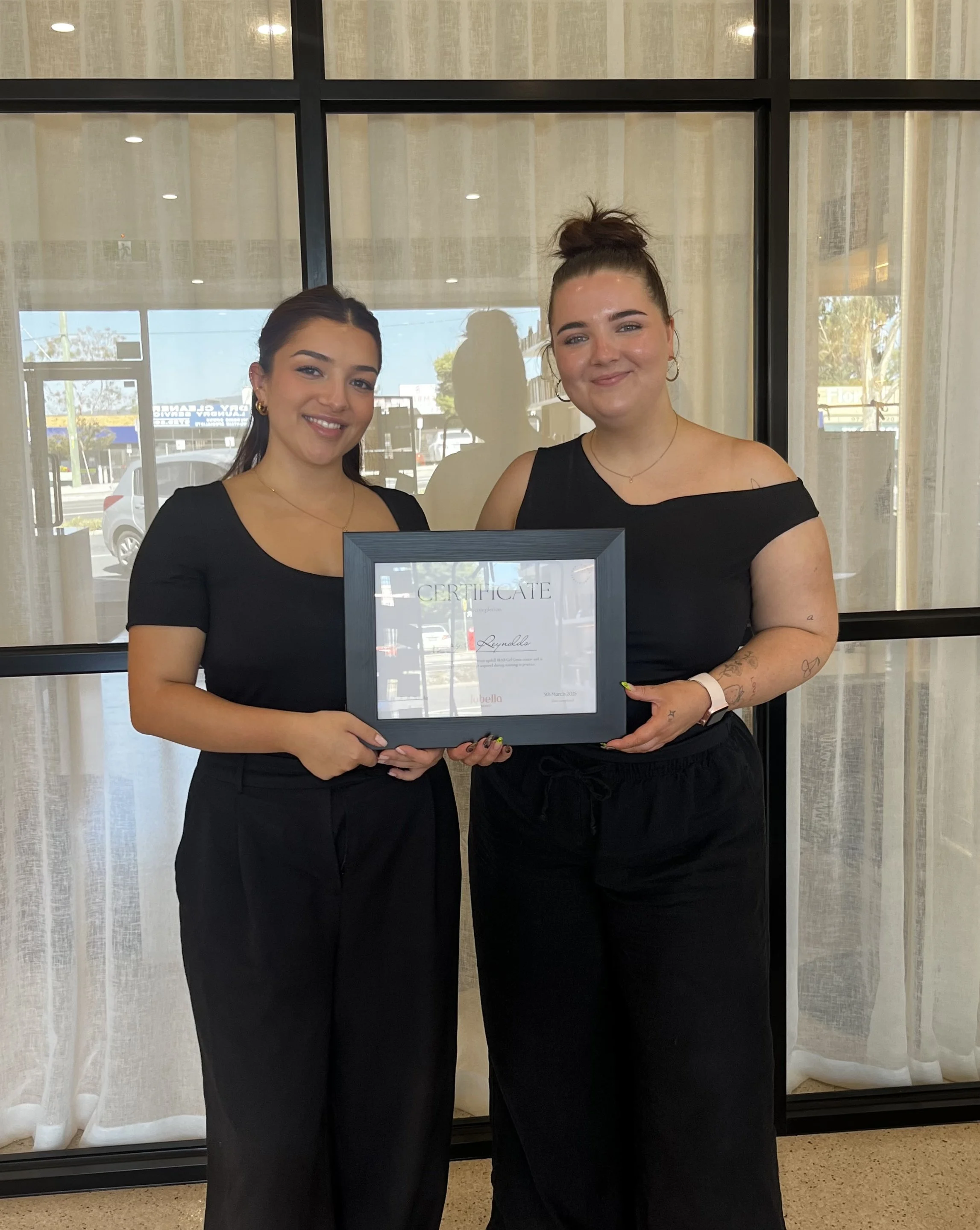 Two women standing together indoors, each wearing black tops and black pants, holding a framed certificate, with a large window behind them.
