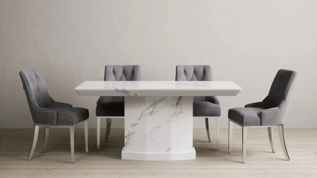 A modern dining room with a white marble table and four gray upholstered chairs with nailhead trim, set against a plain beige wall.