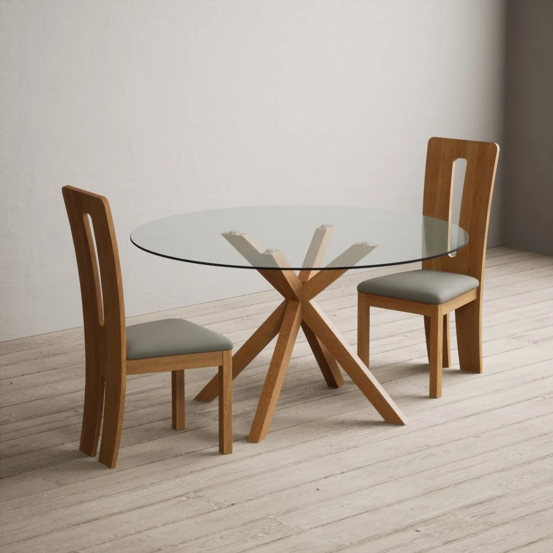 A round glass dining table with a wooden, Y-shaped base, accompanied by two wooden chairs with cushioned seats in a minimally decorated room.