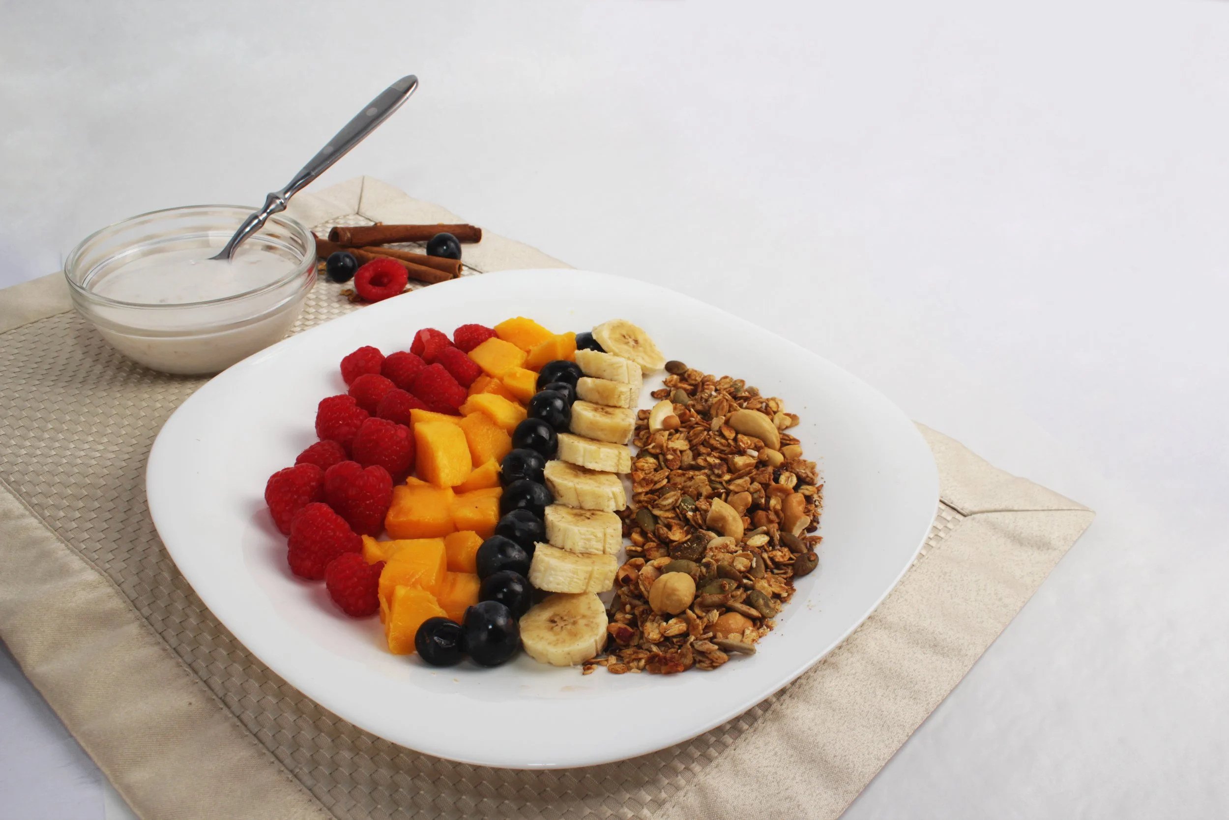 A white plate with sliced bananas, raspberries, mango chunks, blueberries, and granola on a woven placemat.