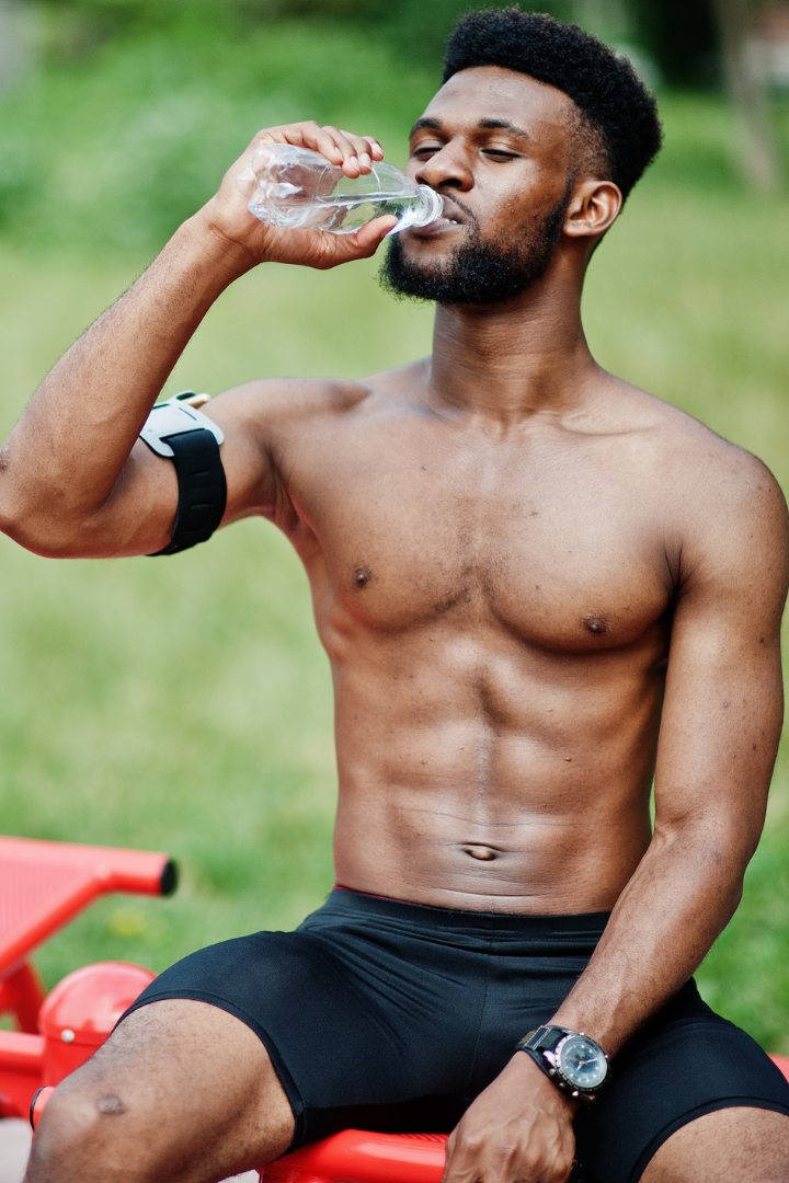 A shirtless young man with a beard and short curly hair drinking water from a plastic bottle outdoors, sitting on a red bench, with a grassy background.