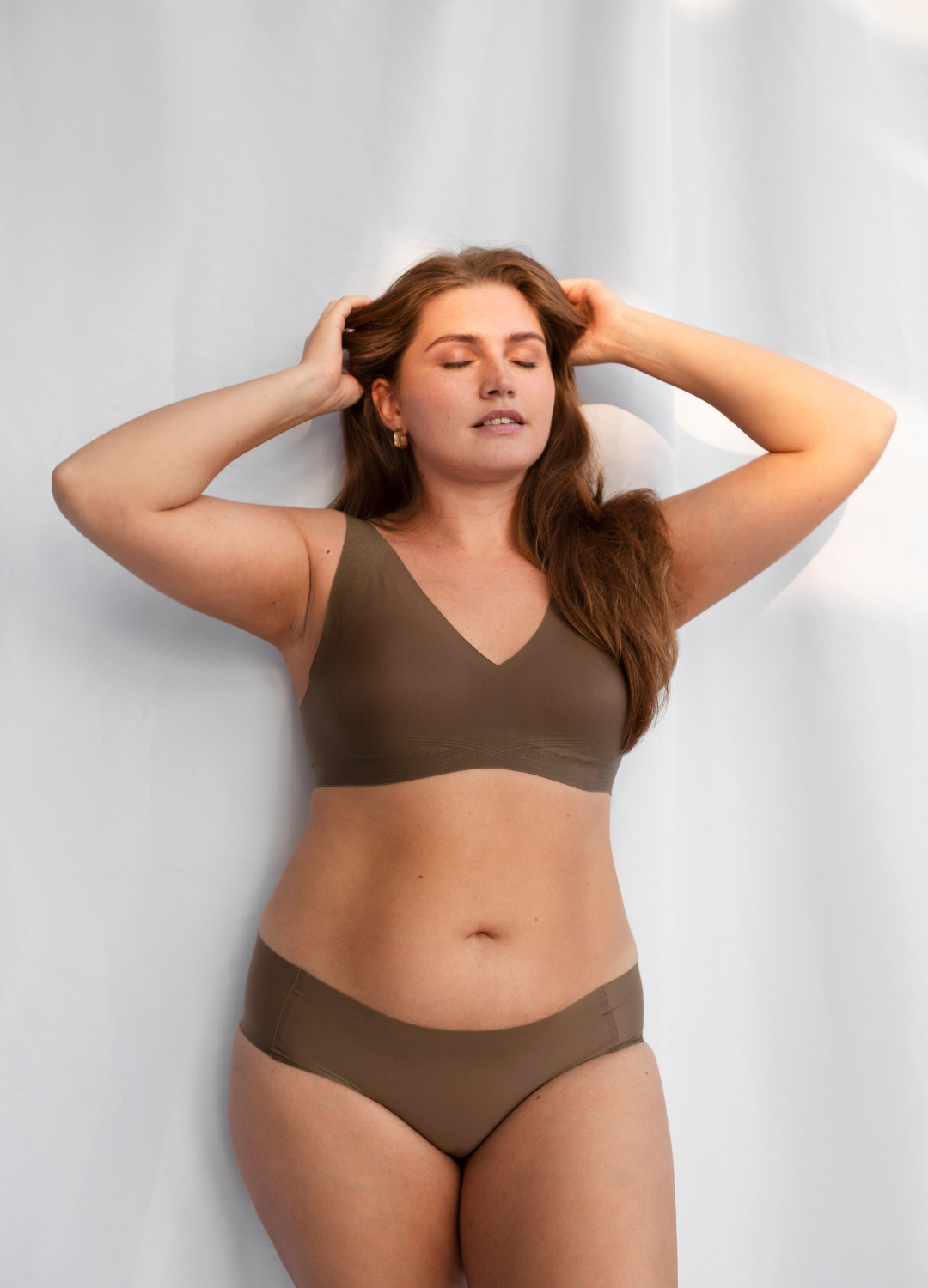 A woman with long brown hair, closed eyes, and a relaxed expression standing against a plain white background. She is wearing a matching olive green bra and bikini-style underwear, with her hands resting on her head and elbows bent.