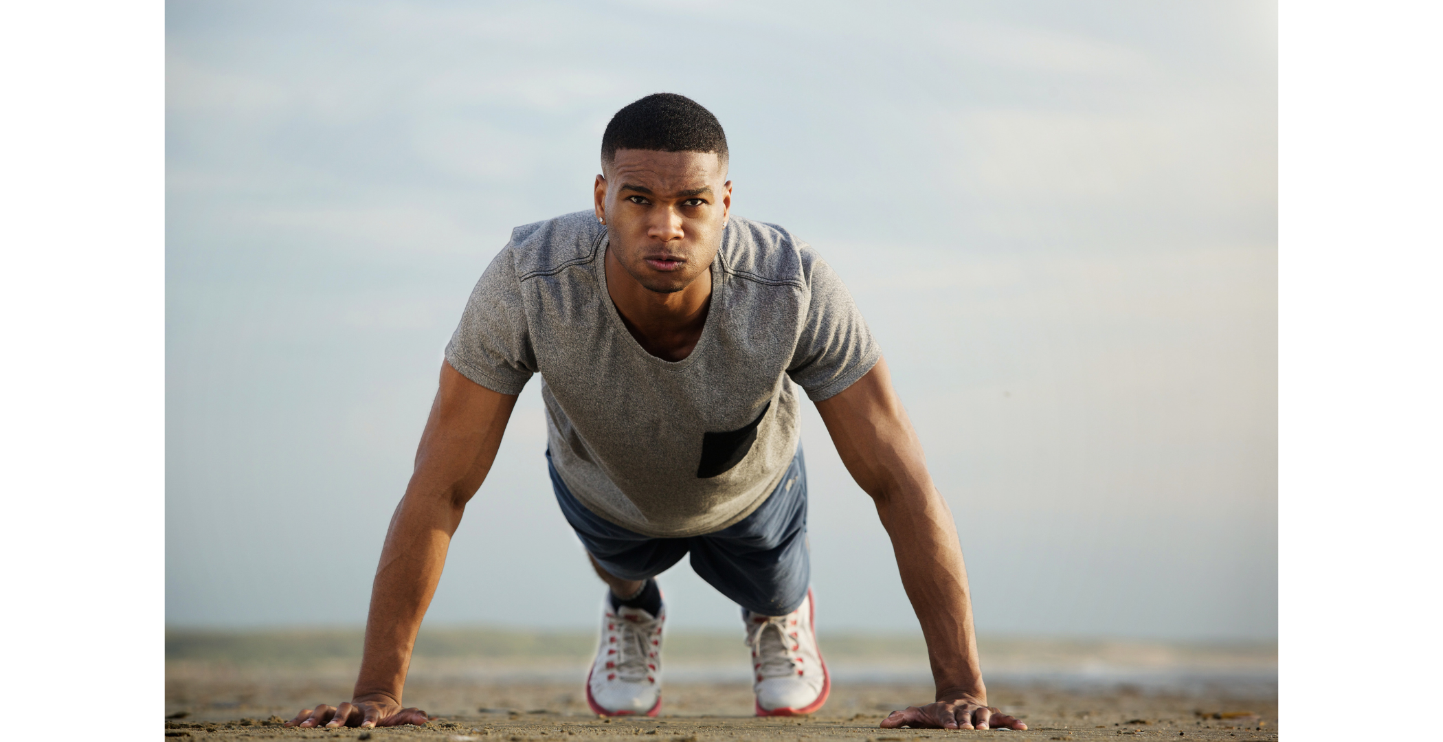 A young man in a gray t-shirt and dark shorts doing push-ups outdoors on a sandy surface, looking into the camera with a serious expression.