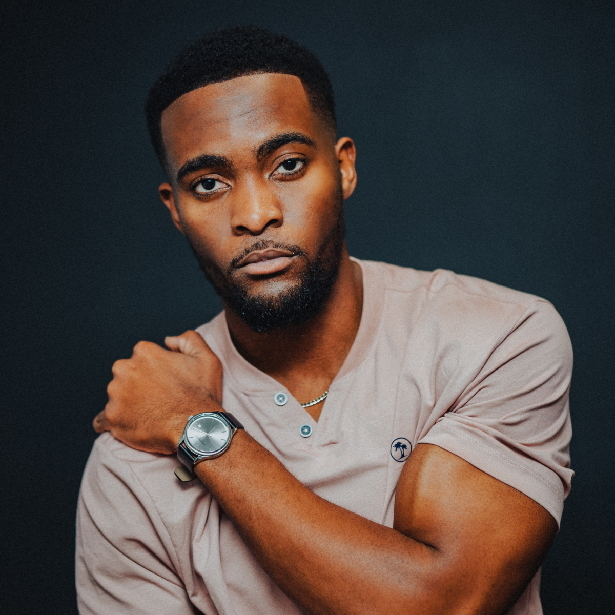 A young man with a beard and short hair posing against a plain dark background, wearing a light-colored t-shirt and a watch on his left wrist.