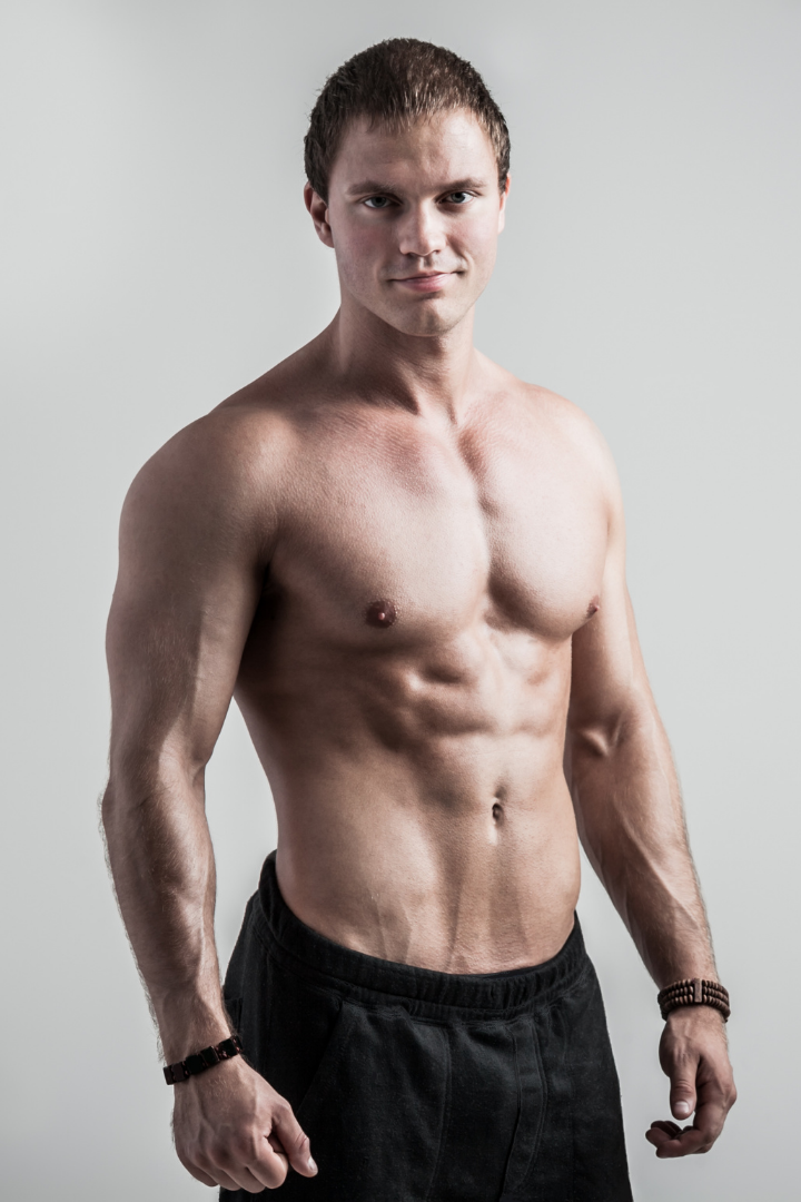 A shirtless young man with short brown hair and a lean, muscular physique, wearing black pants and bracelets, standing against a plain gray background.