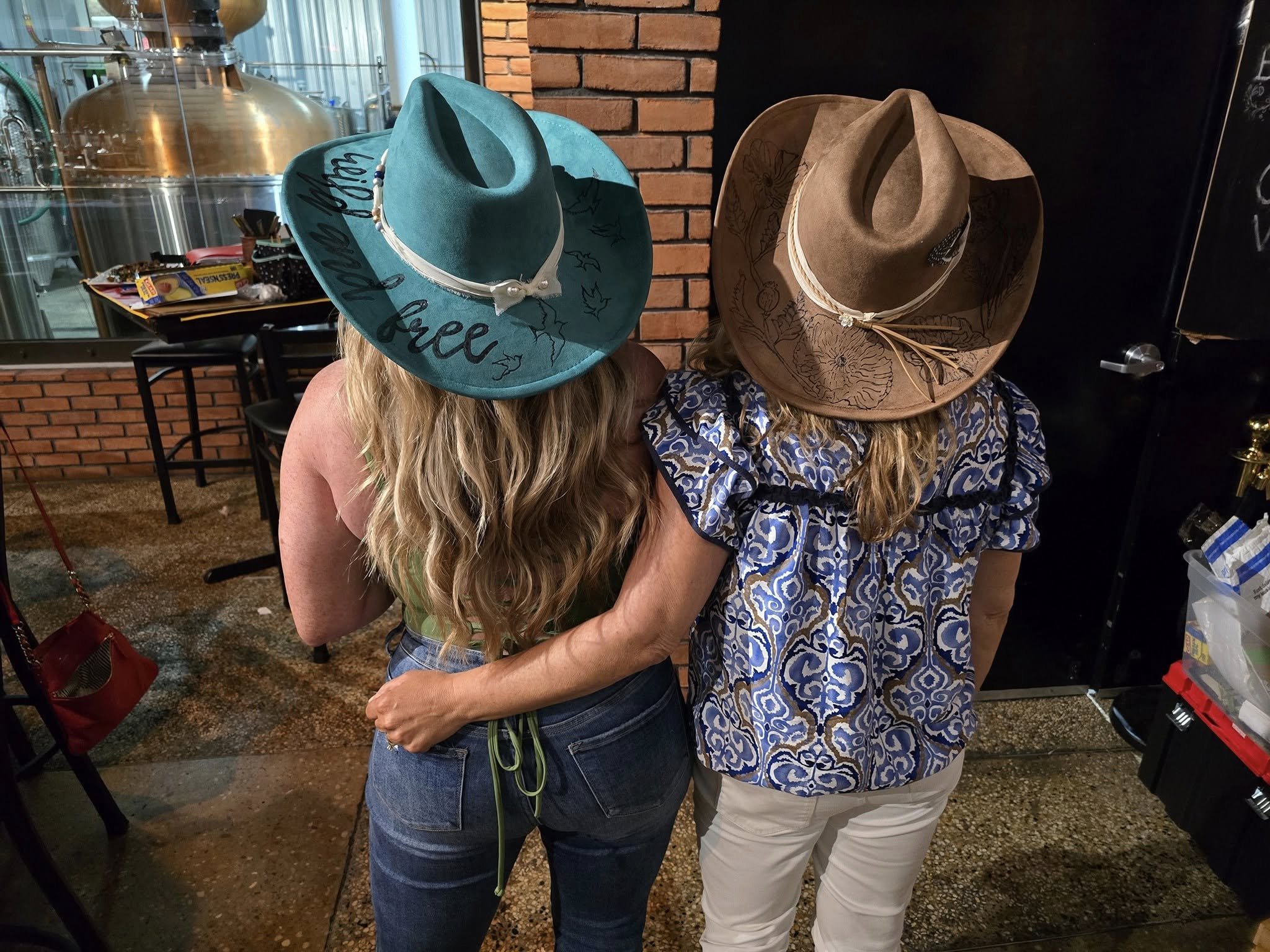 Two women wearing cowboy hats hugging each other inside a brewery or bar with brick wall and brewing equipment in the background.