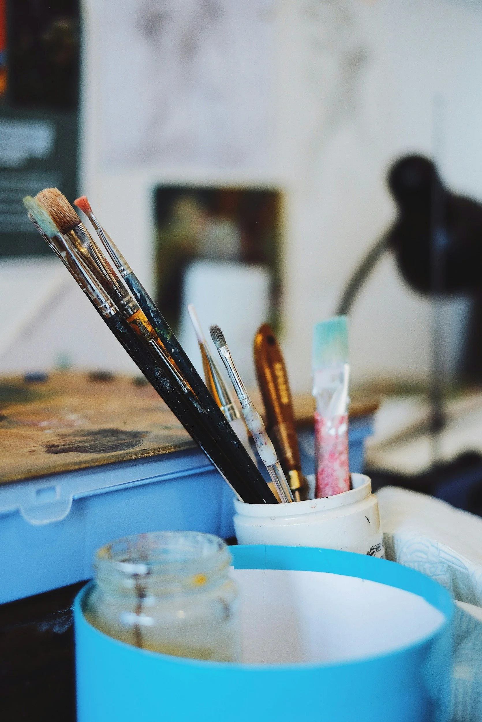 Close-up of paintbrushes stored in a white container on a cluttered workspace with paint jars and supplies.