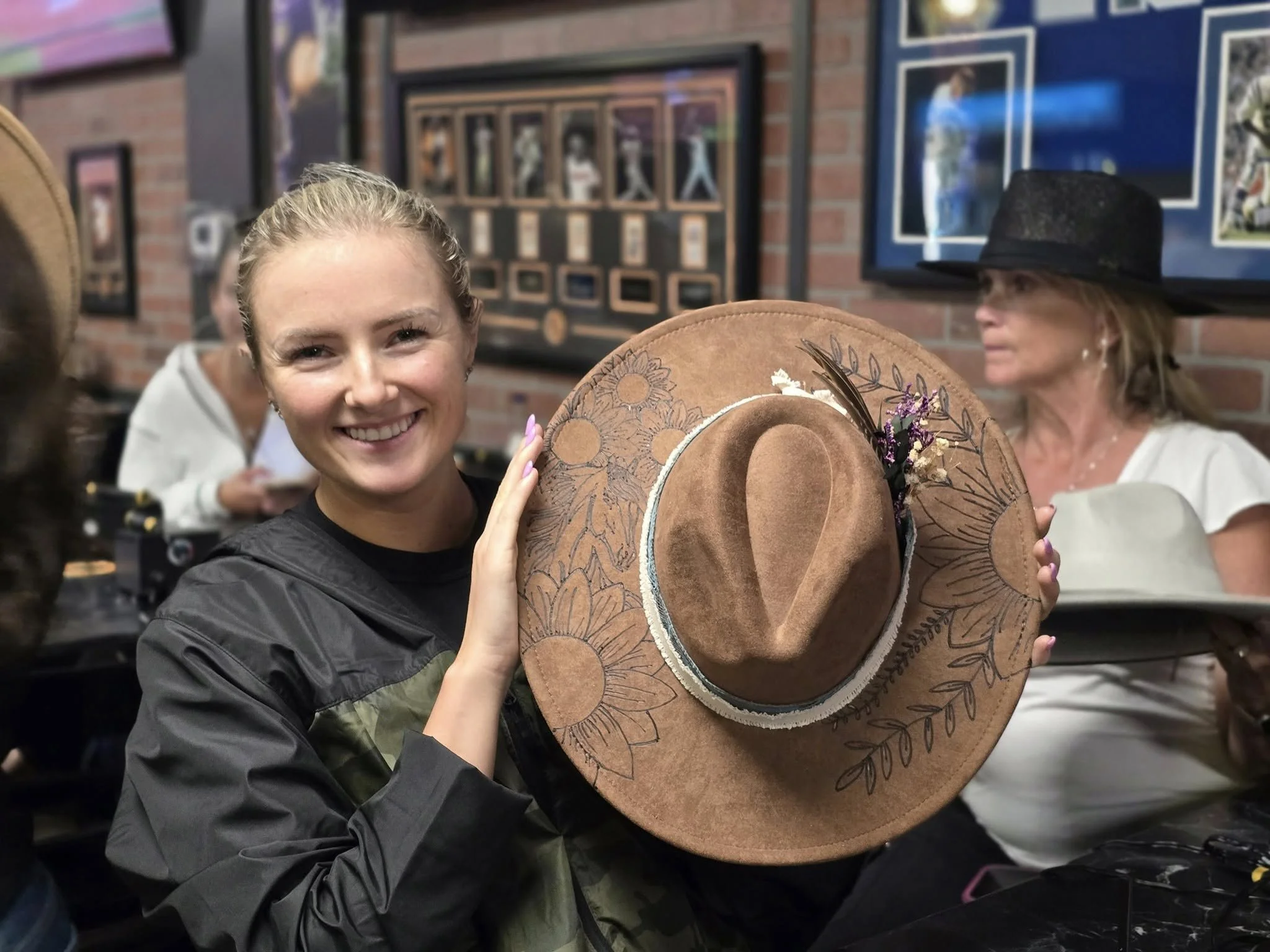 A woman smiling while holding a large, decorated cowboy hat in front of her face in a room with framed photos on the wall.