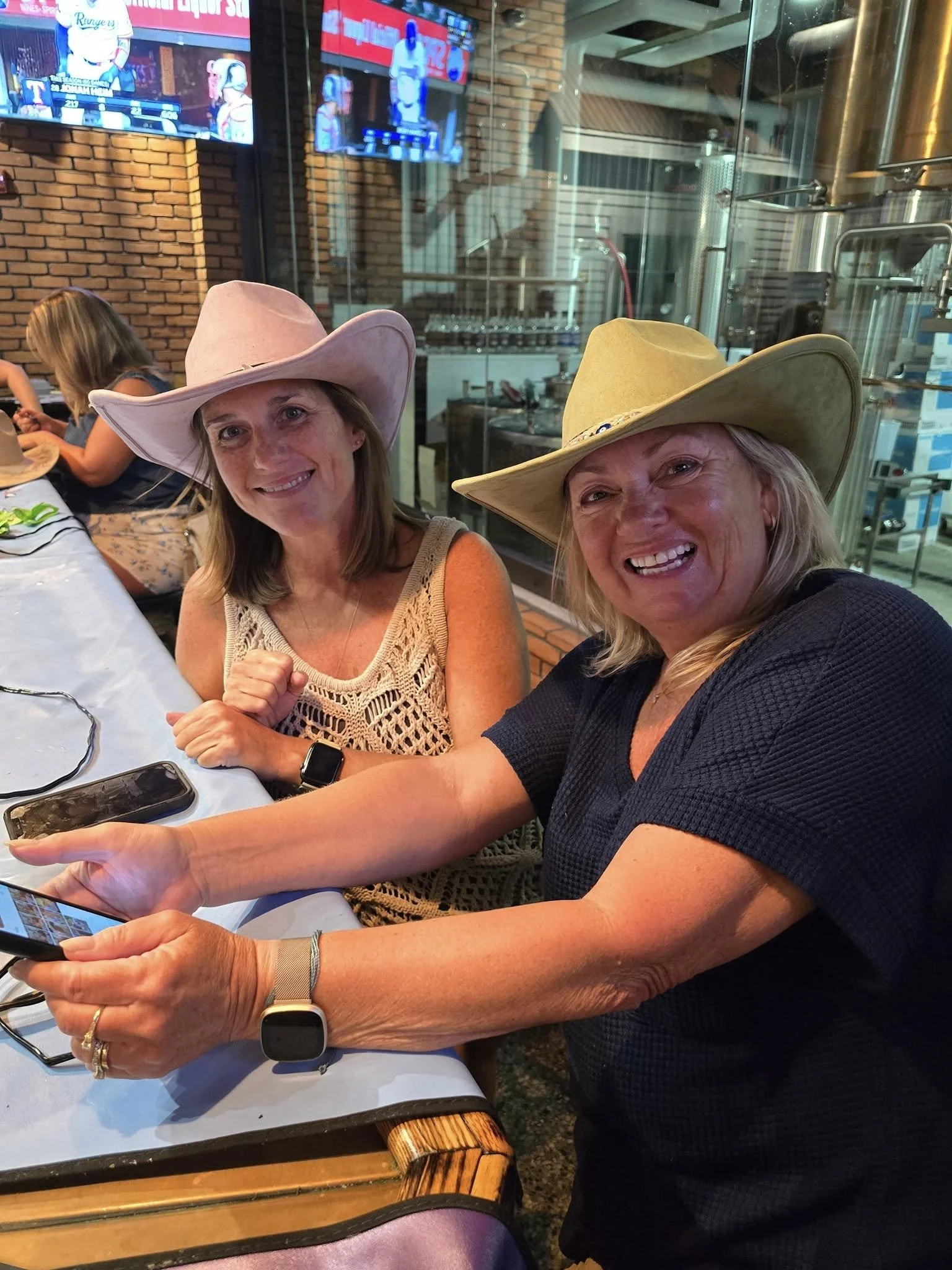 Two women sitting at a table, wearing large cowboy hats, taking a selfie in a restaurant or bar with a brick wall and TV screens in the background. One woman is smiling and the other is looking at her phone.