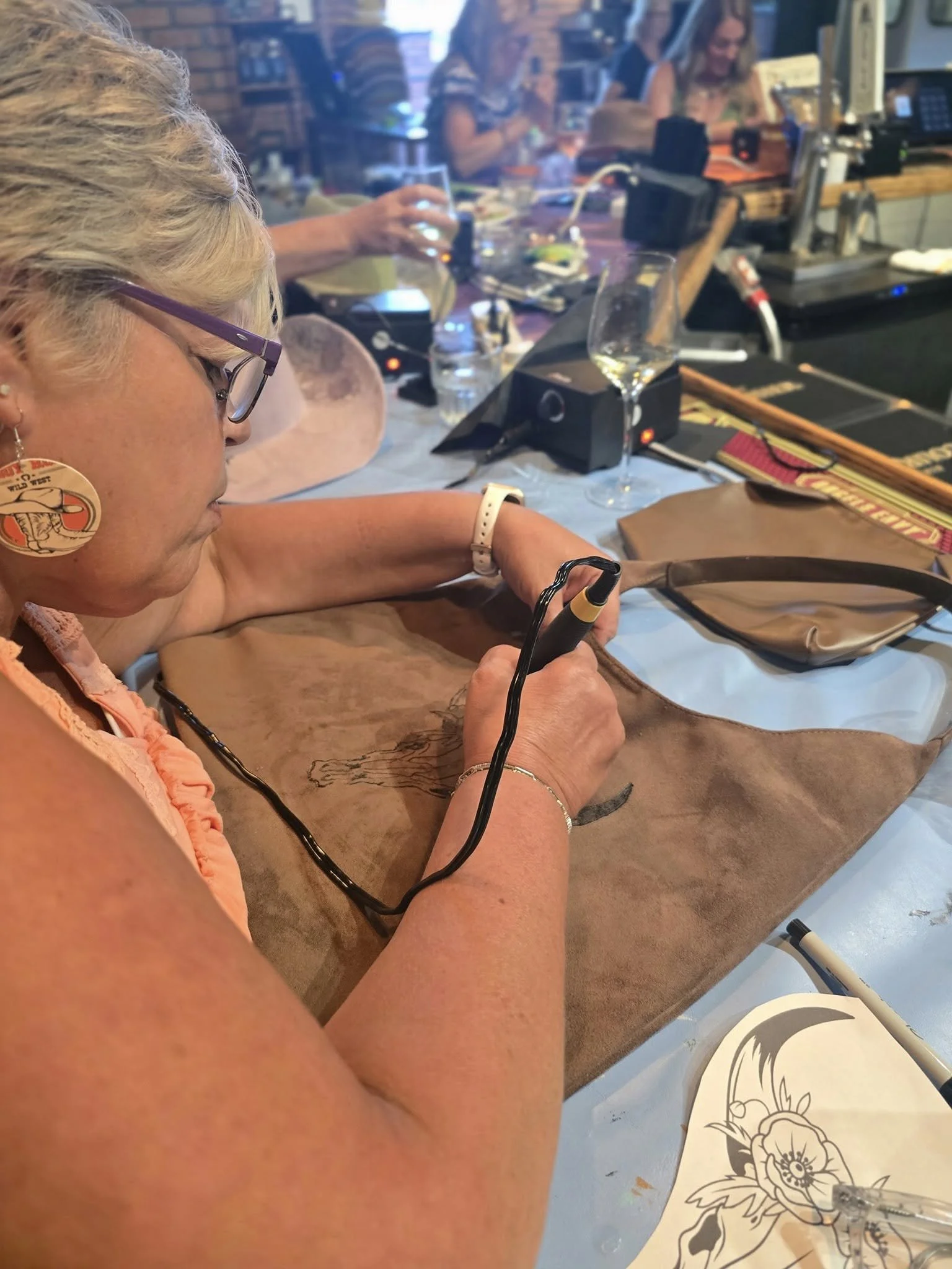 A woman with short blonde hair, glasses, and earrings sitting at a table with wood burning tool and a bag burn design in progress, with other people in the background in a restaurant.