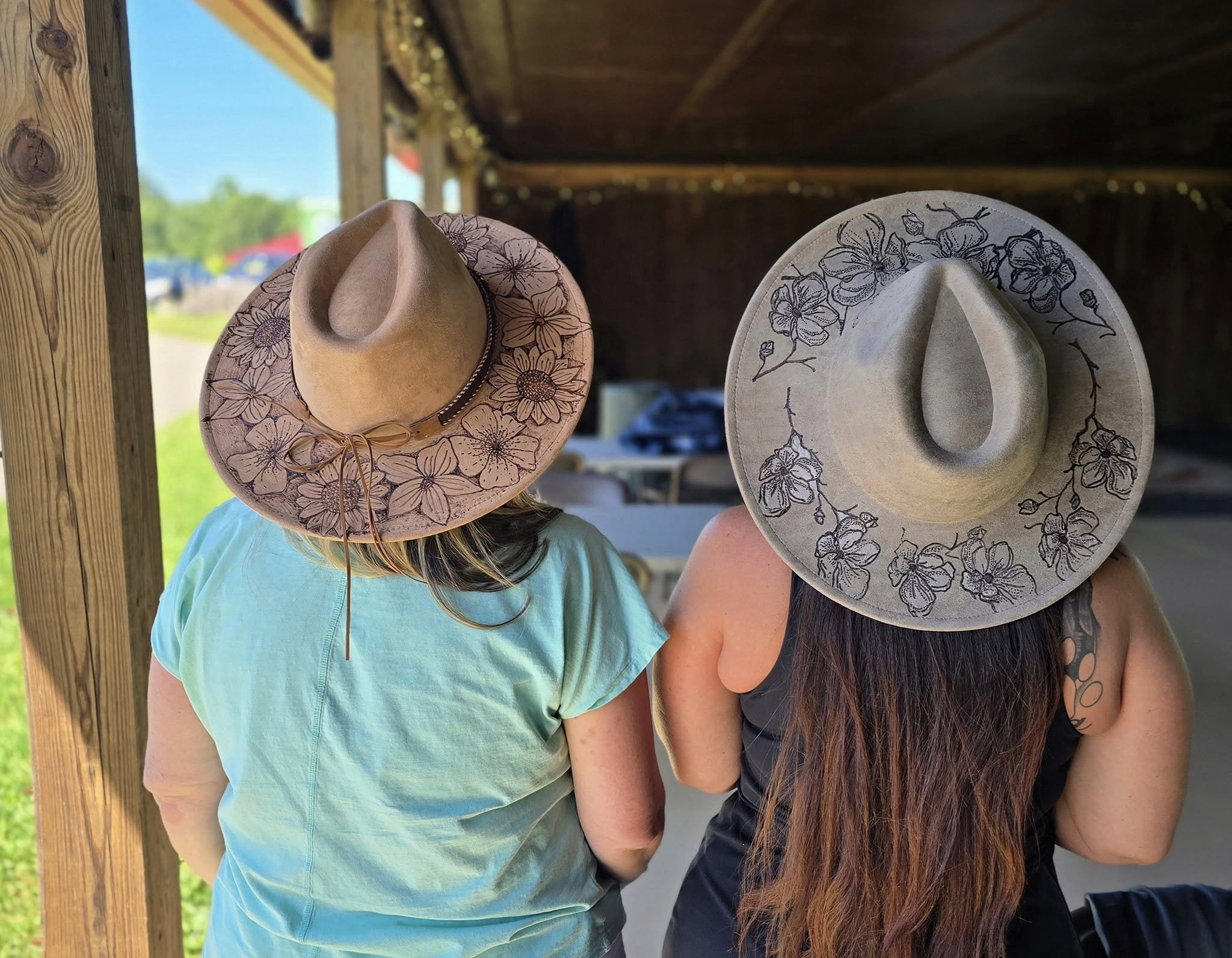 Two women with long hair wearing wide cowboy straw hats decorated with floral burn patterns, standing close together outdoors near wooden posts.