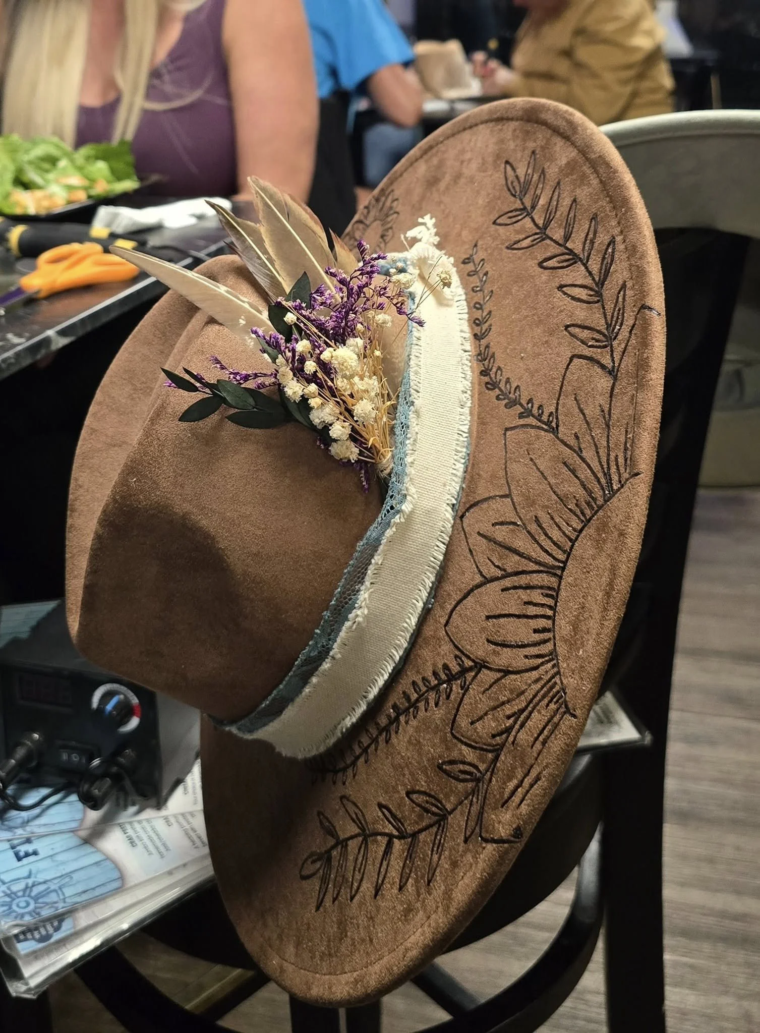 Close-up of a brown fedora hat with black leaf pattern burnt into the hat brim, decorated with a small bundle of dried flowers and feathers.