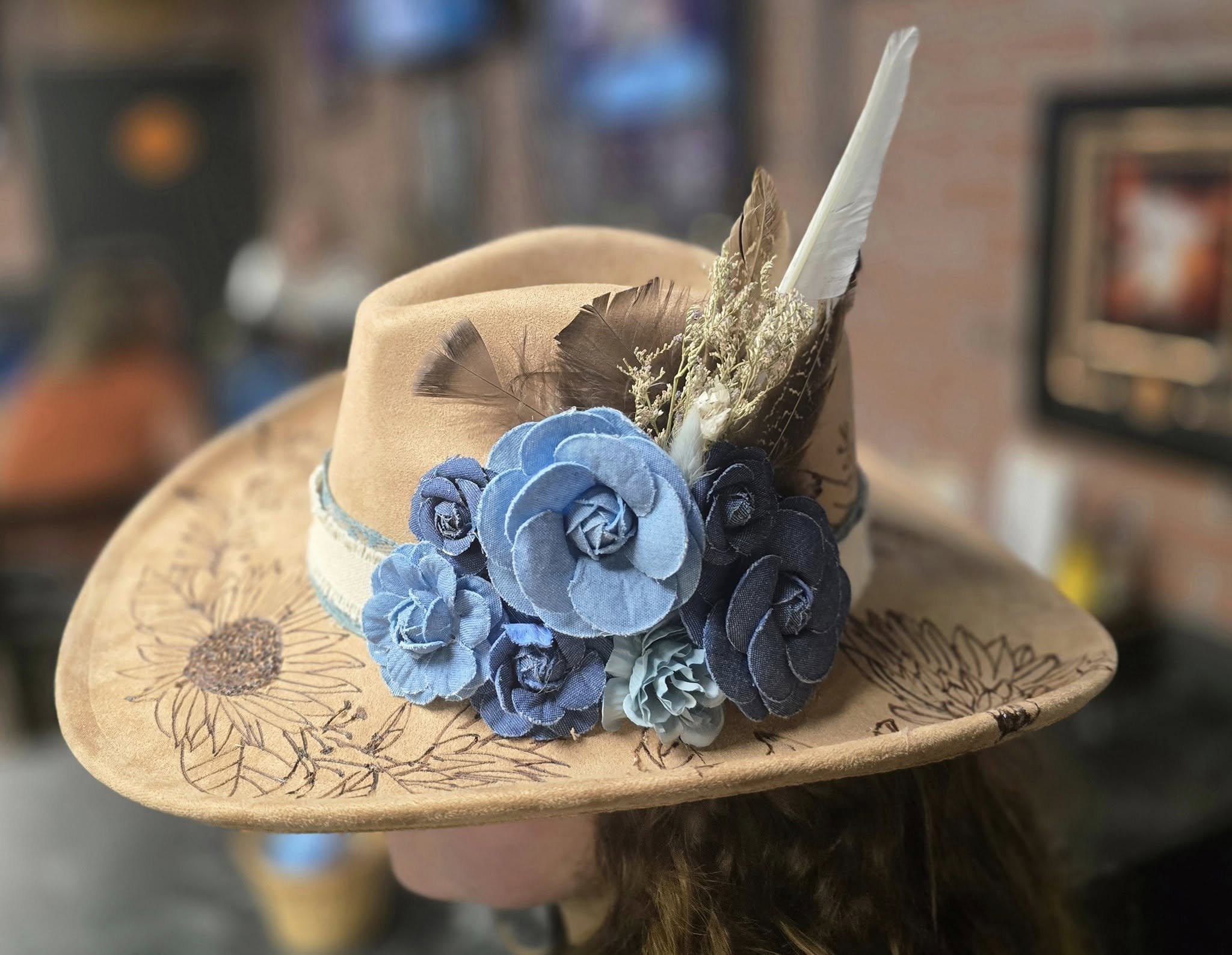A tan hat with a burnt floral pattern, decorated with blue and brown feathers, resting on a wooden surface.
