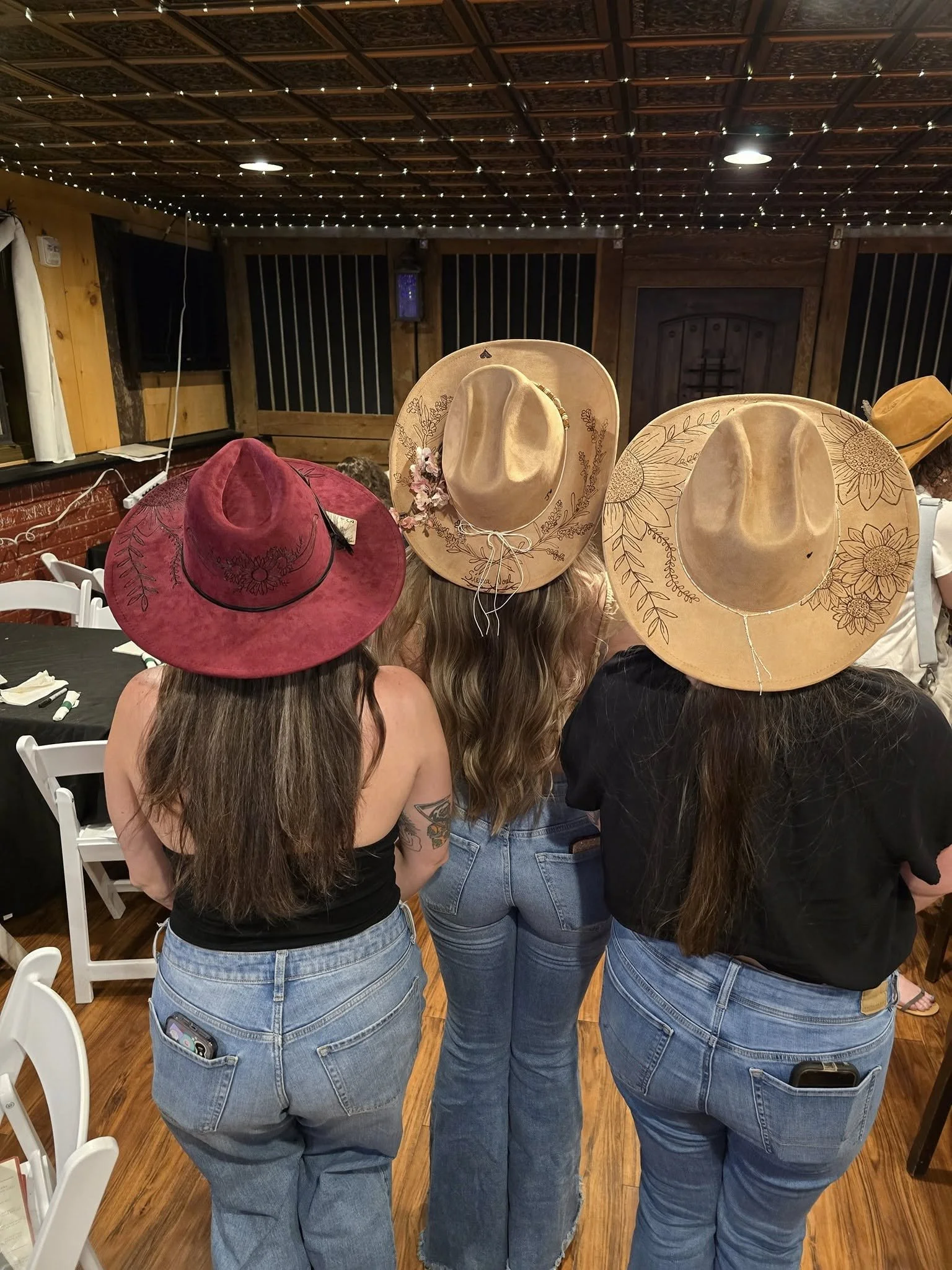 Three women standing together wearing large cowboy hats, taken from behind. The women are dressed in black, tank tops, and jeans inside a wooden decorated room with string lights on the ceiling.