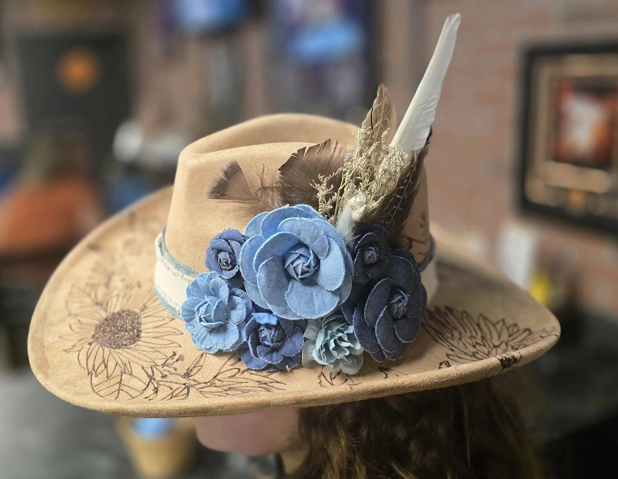 A hat decorated with blue fabric flowers and feathers. A hand burnt design on the hat.