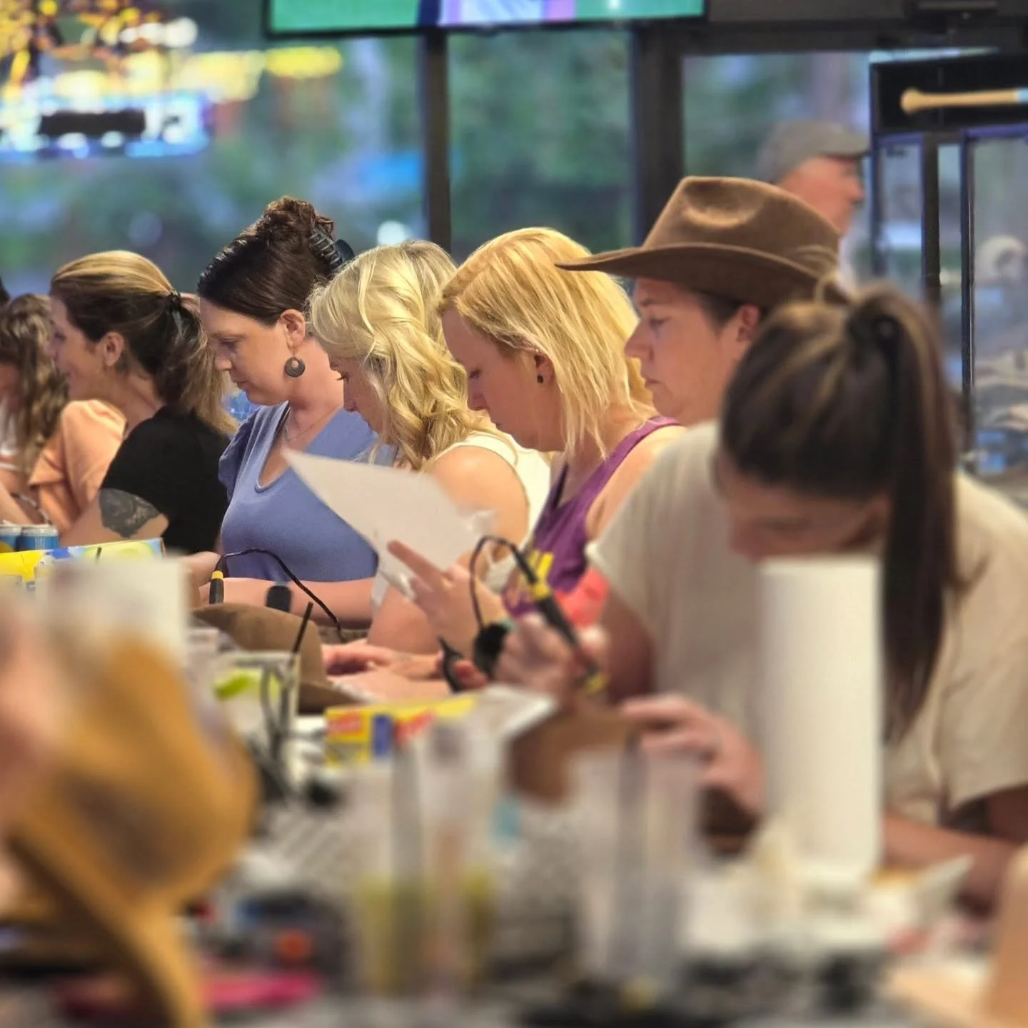 People seated at a table at a hat burning workshop, engaging in fun conversations and creating custom hats and bags under the direction of Claudia's Traveling Palette.