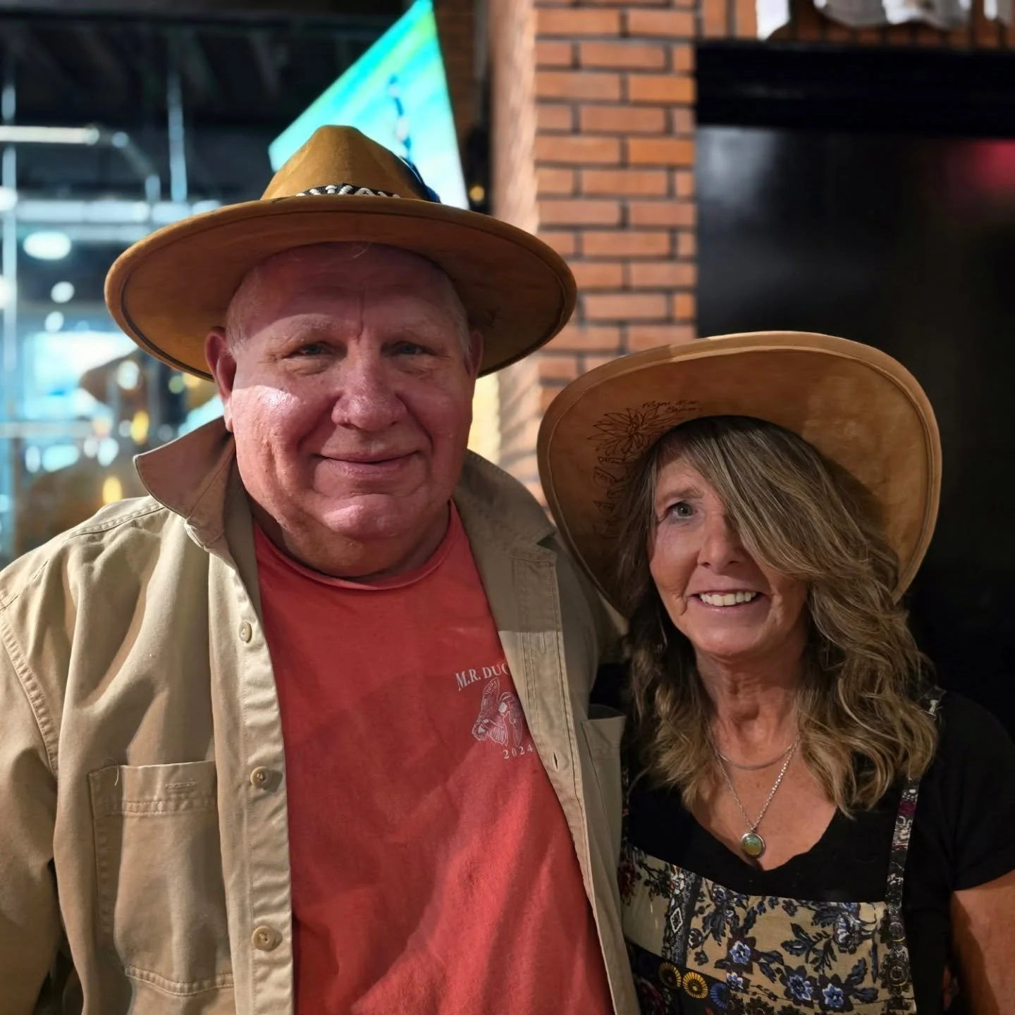 A smiling man and woman wearing cowboy hats inside a restaurant, with brick walls and a television screen in the background.