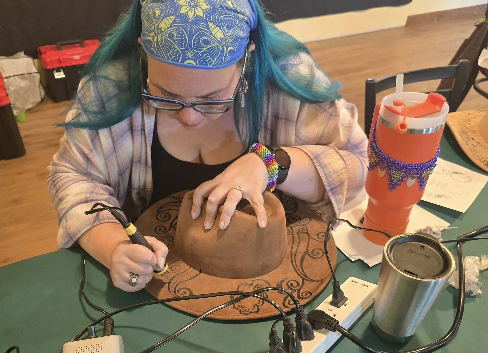 A woman with turquoise hair, wearing glasses and a blue headscarf with yellow butterfly patterns, is sitting at a table using a wood burner to create a custom design on a cowboy hat.