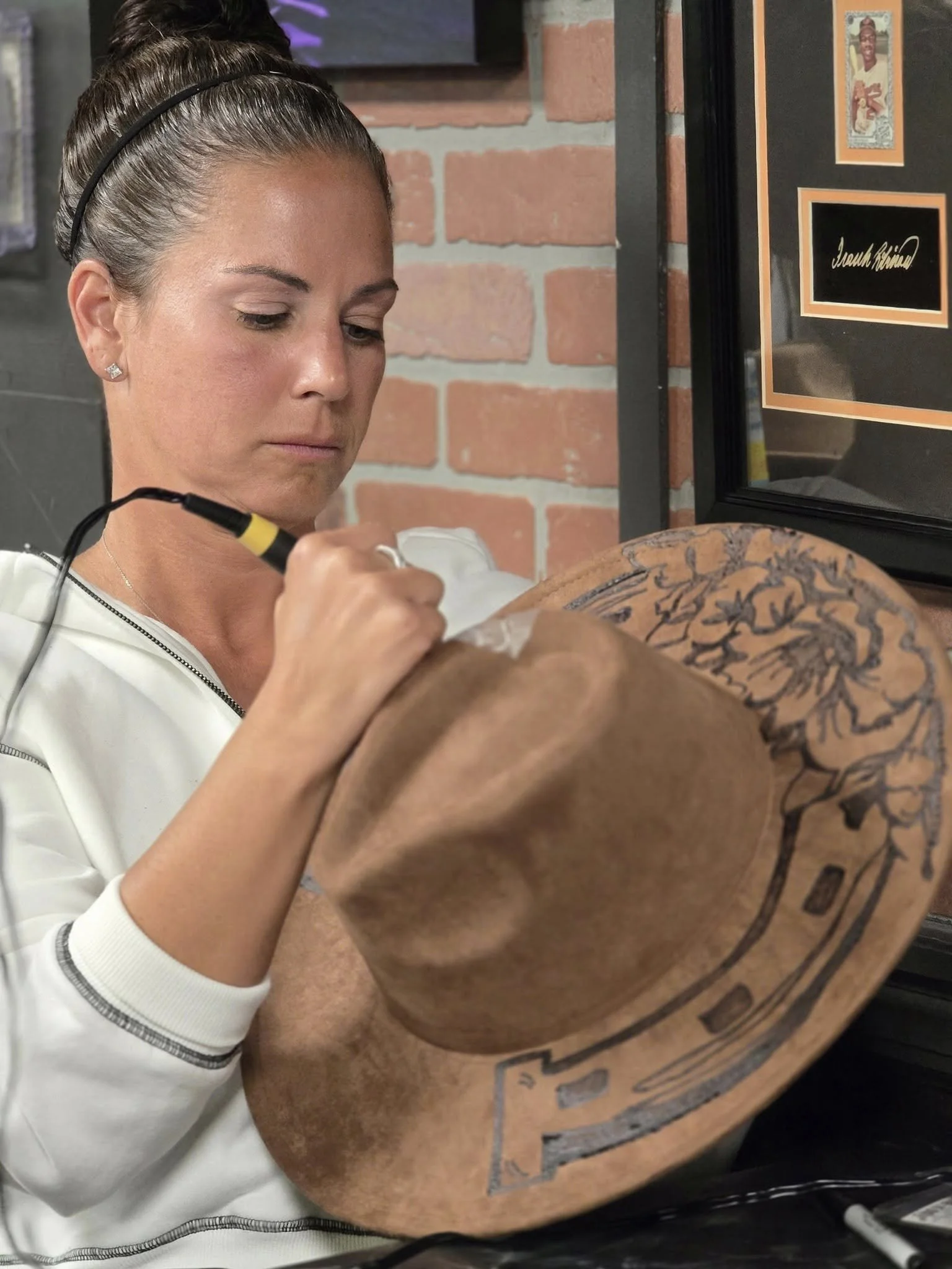 A woman with brown hair tied in a bun, wearing earrings and a white top, burning a custom design into a cowboy hat using a woodburner.