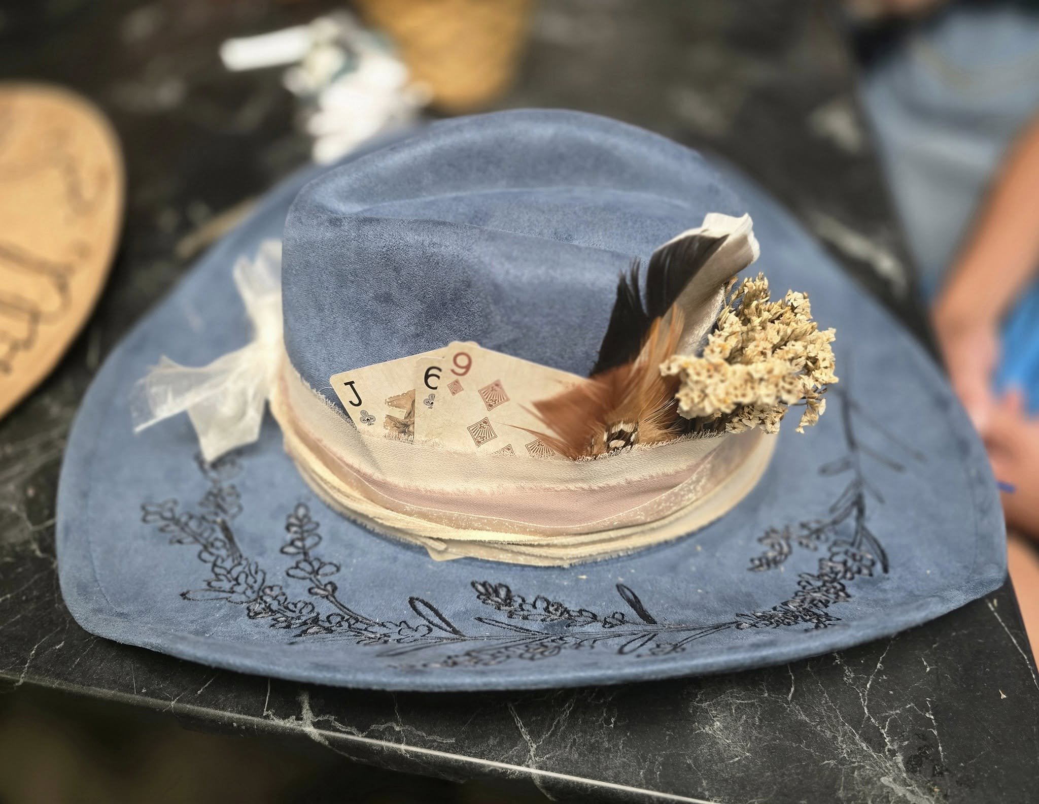 A cowboy hat decorated with a deck of playing cards, including the Jack, 6, and 9 of spades, dried herbs or flowers, on a black table.