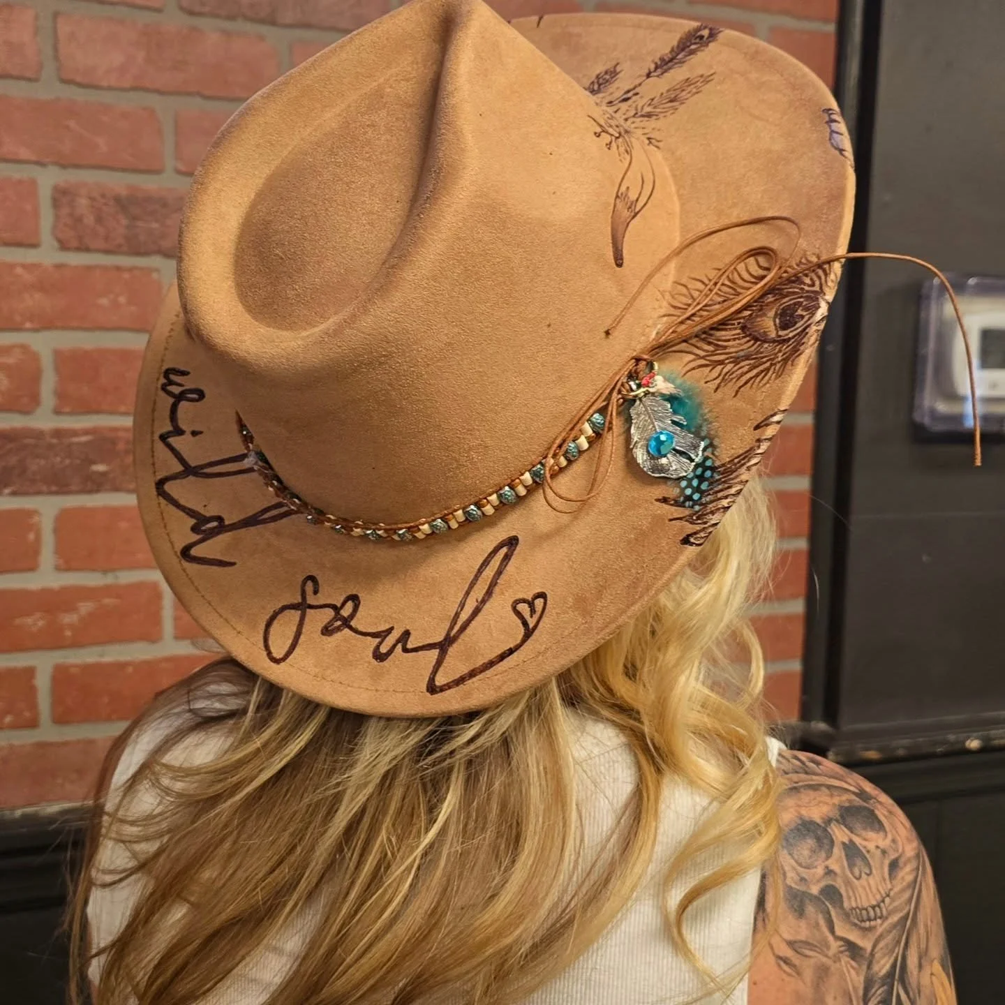 A tan cowboy hat decorated with beads, writing, and artwork, worn by a person with long blonde curly hair and a tattoo of a skull on their left arm, against a brick wall background.