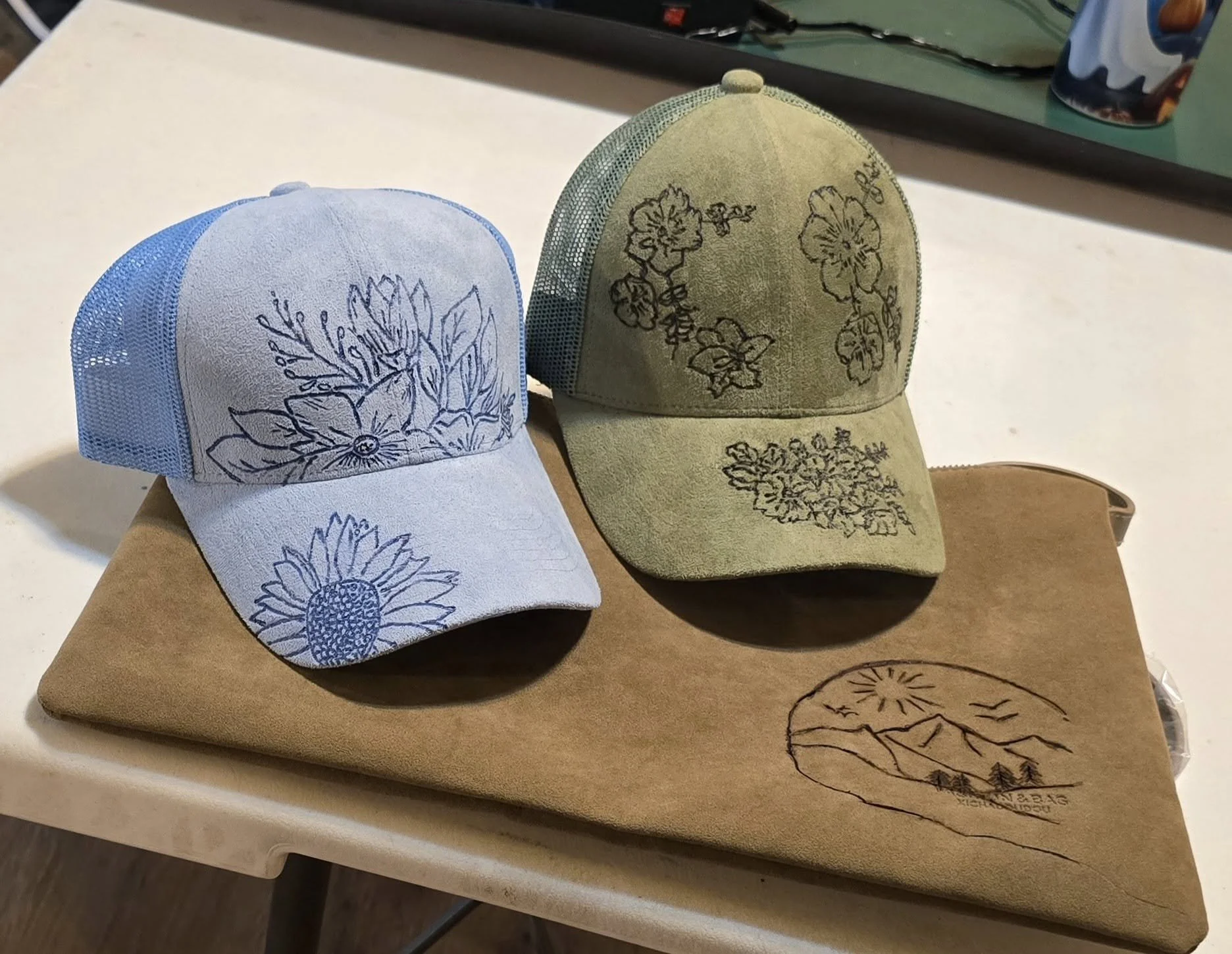 Two baseball caps, one light blue and one beige, both with black hand-burnt flower illustrations, resting on a folder with a matching logo, on a white surface.