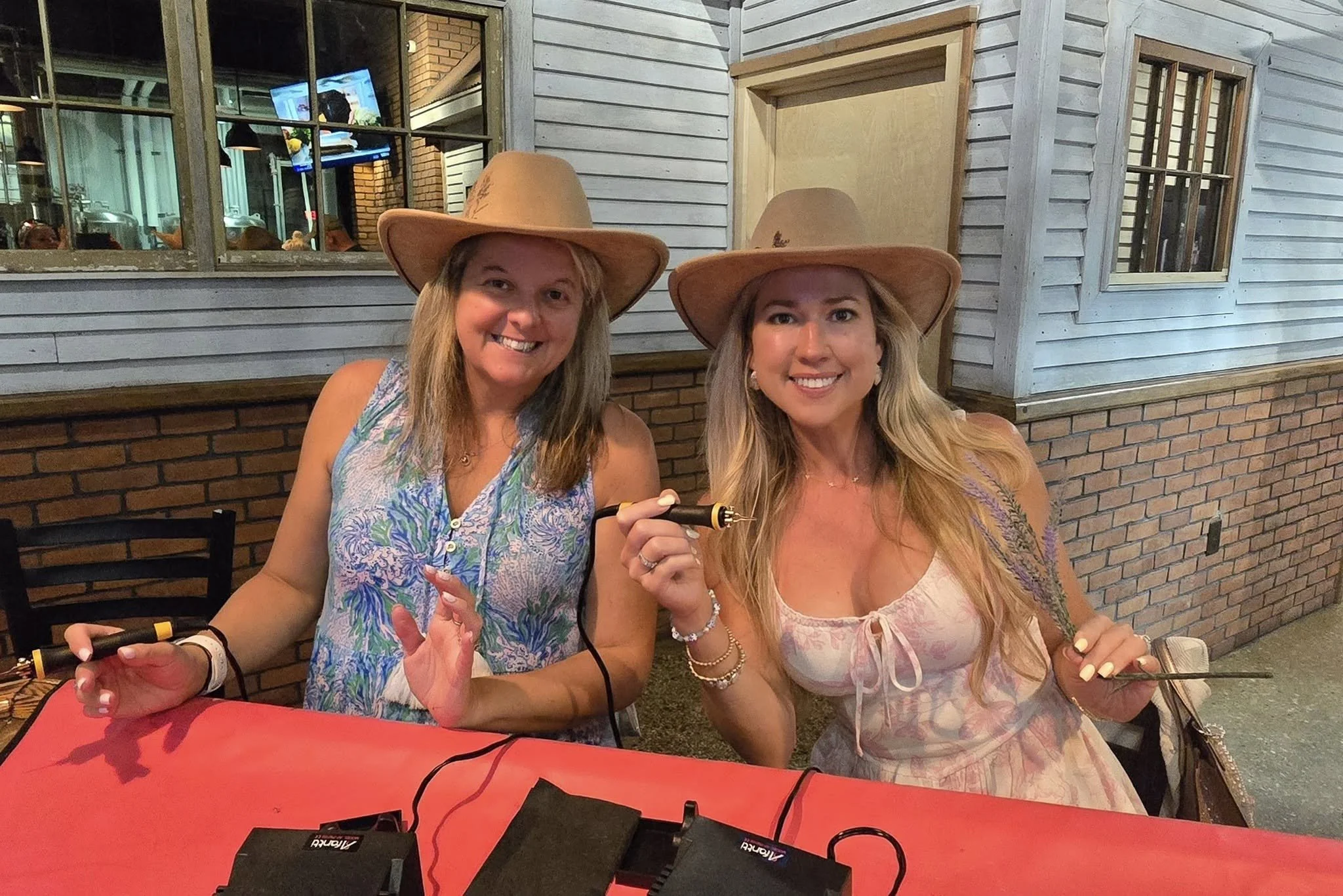 Two women sitting at a table inside a restaurant, both wearing large tan cowboy hats and smiling at the camera. One woman is holding a woodburner and the other has her hand near her face. The background shows wood paneling, windows, and a brick wall.