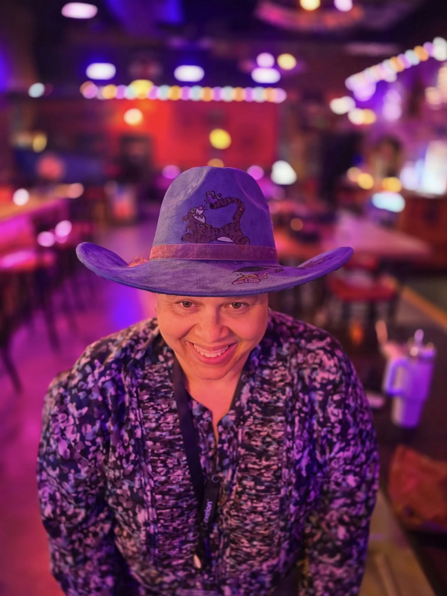 Smiling person wearing a wide-brimmed hat with a tiger embroidery, indoors with colorful, blurred neon or LED lights.