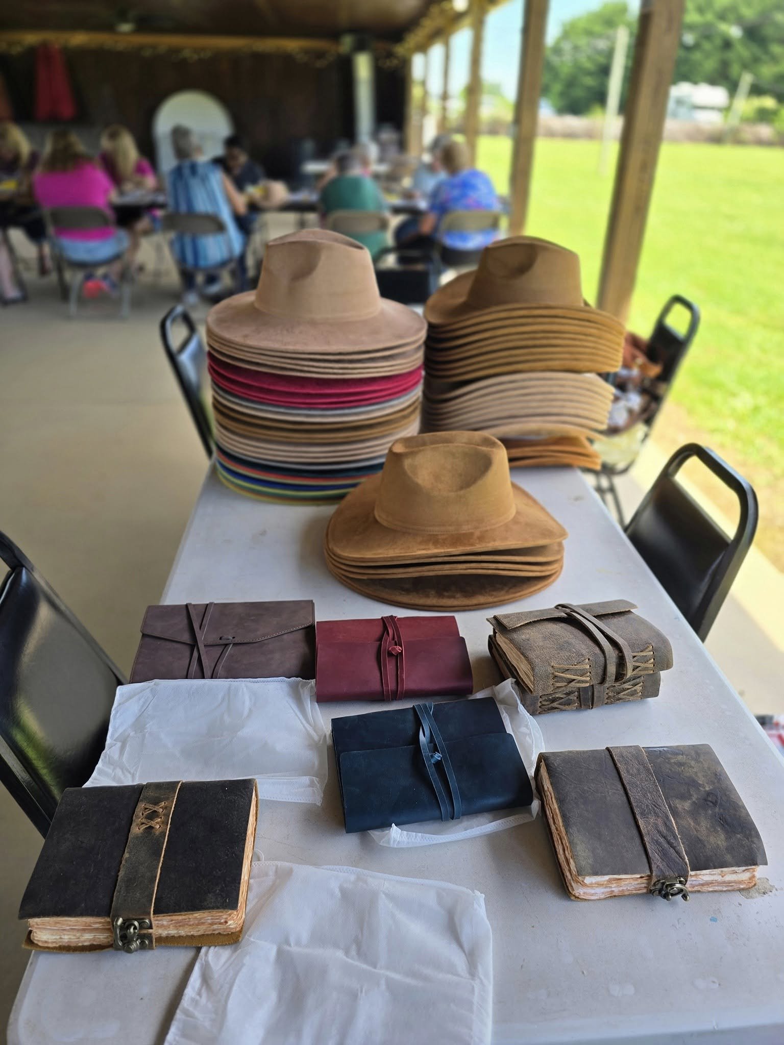Stacks of cowboy hats, leather-bound journals, and wrapped notebooks on a white table, with blurred people sitting at tables in an outdoor covered pavilion and green grass in the background.