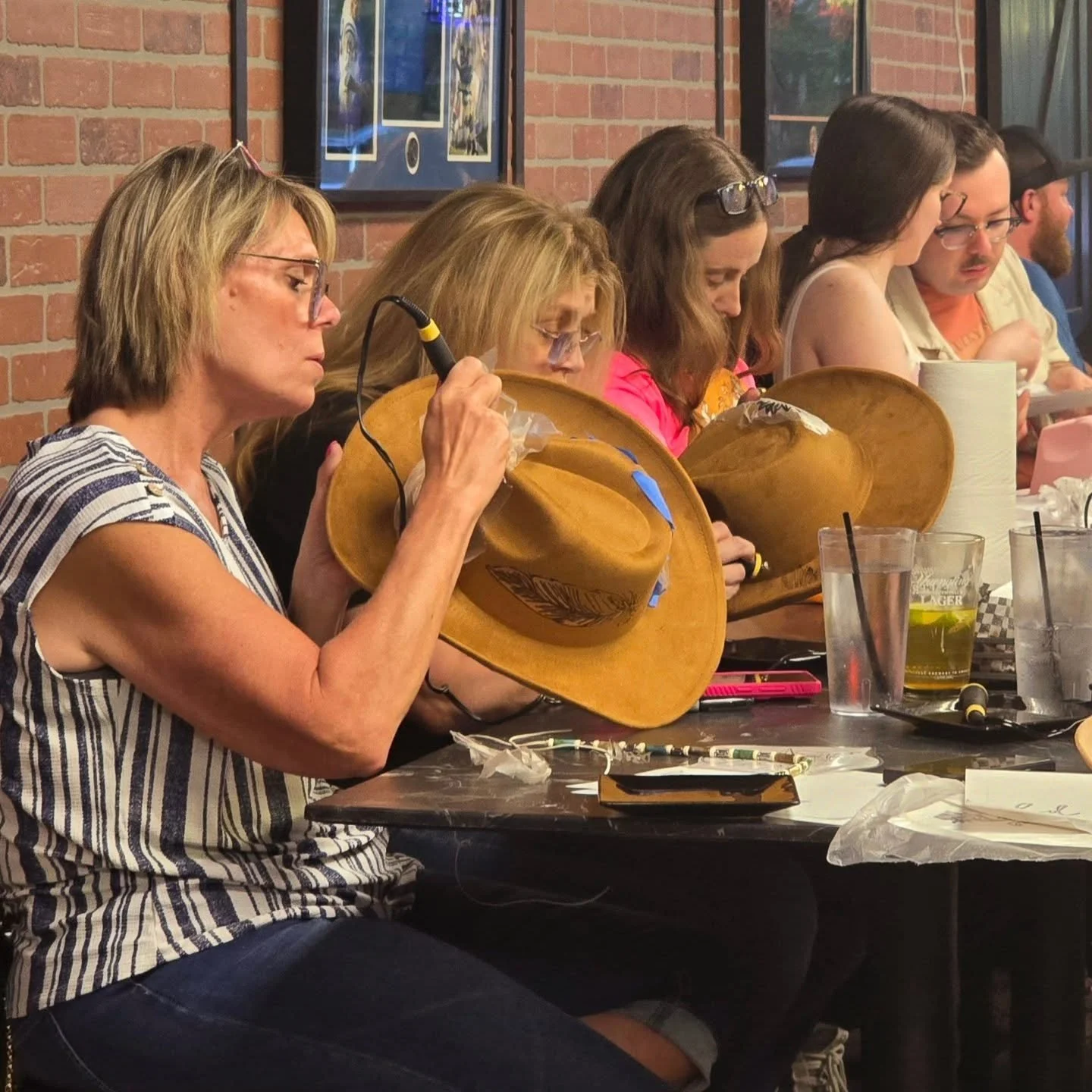 Group of people sitting at a restaurant table wearing cowboy hats, with drinks and miscellaneous items on the table, against a brick wall background.