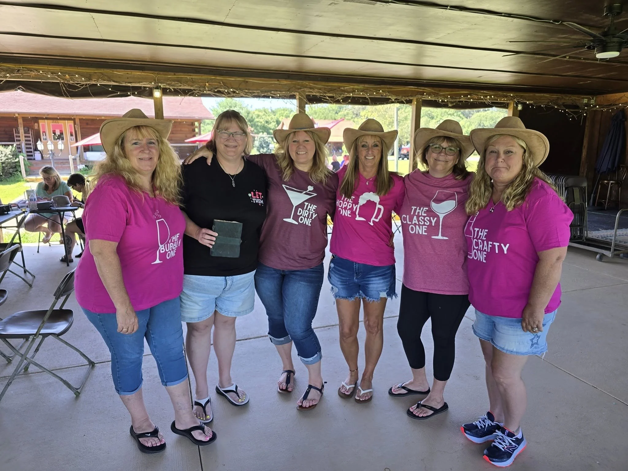 Six women standing closely together at an outdoor event, wearing matching bright pink t-shirts and wide-brimmed hats, smiling for a group photo.