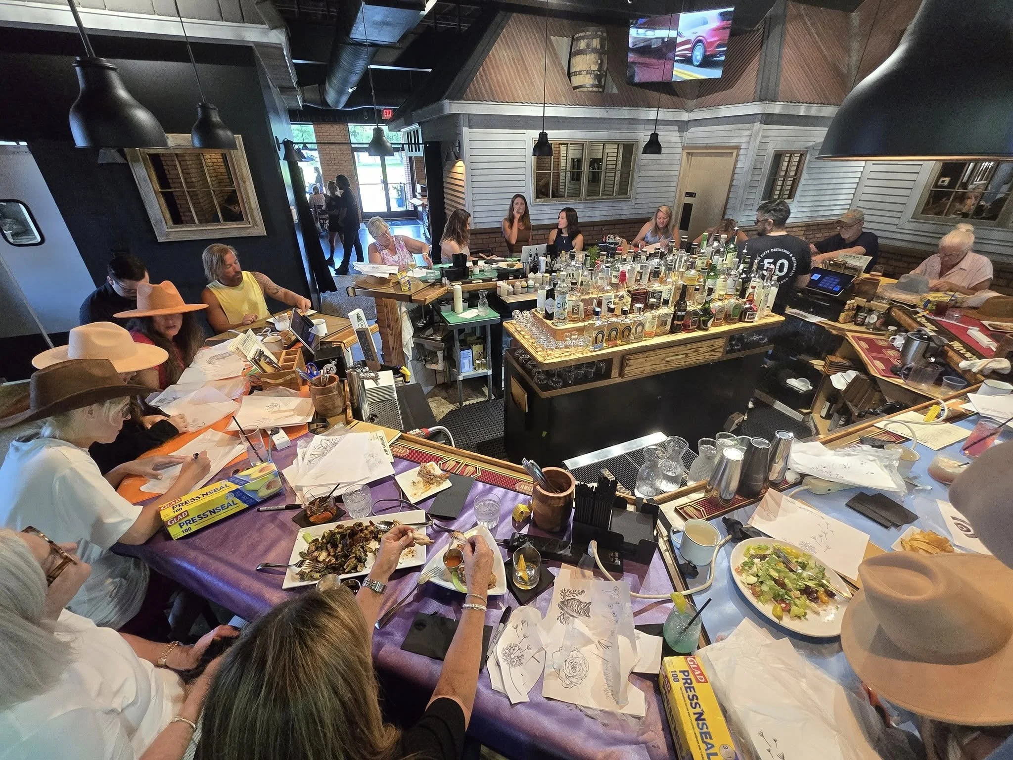 A group of people sitting around a table with food and drinks in a restaurant or bar. There is a bar area with various bottles of alcohol in the center of the image. All people are at a hat burning workshop and creating custom hats and bags.