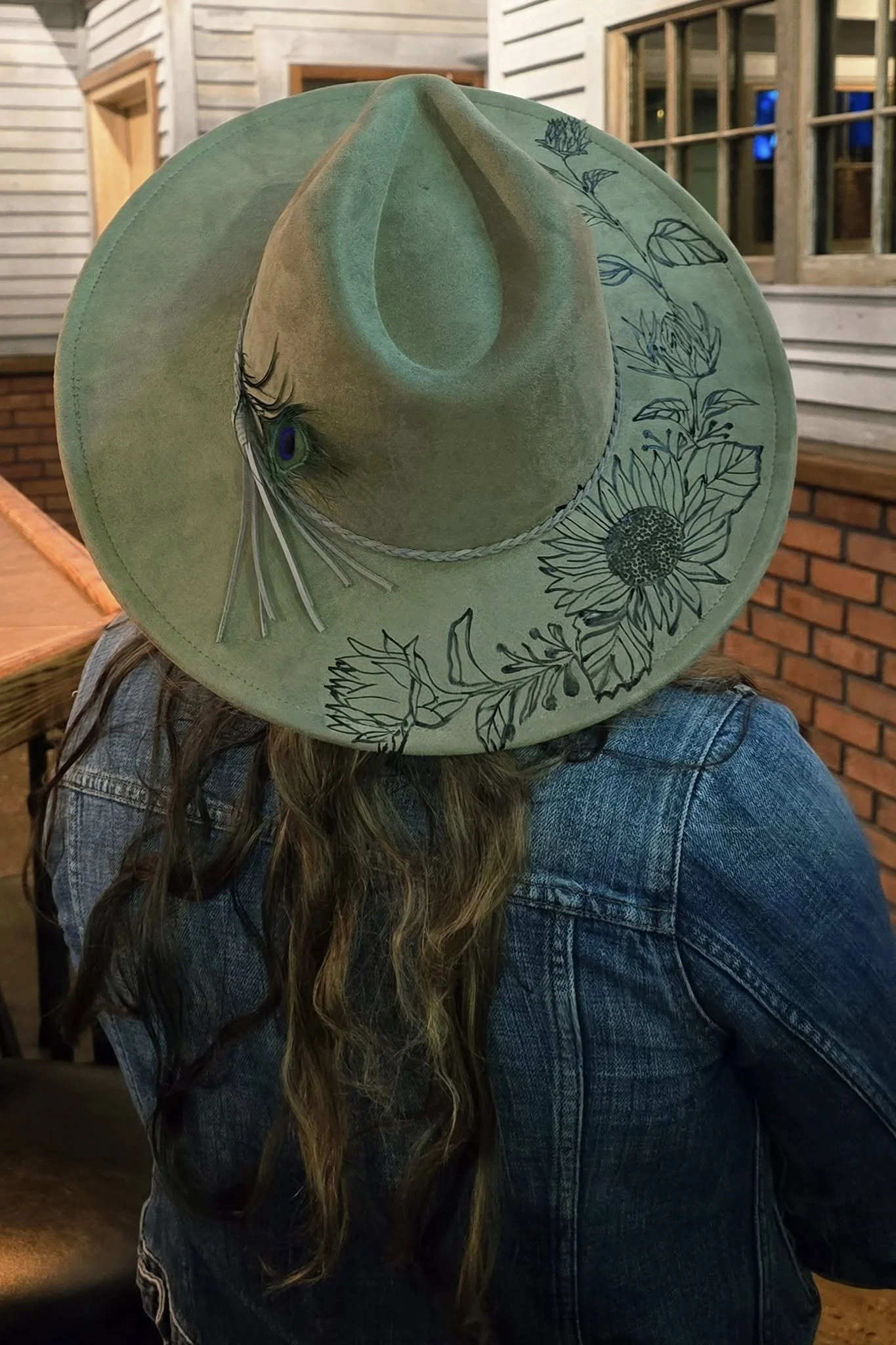 A person wearing a green cowboy hat with burnt floral and peacock feather design, a braid detail, and long hair, sitting indoors with wooden walls and a window