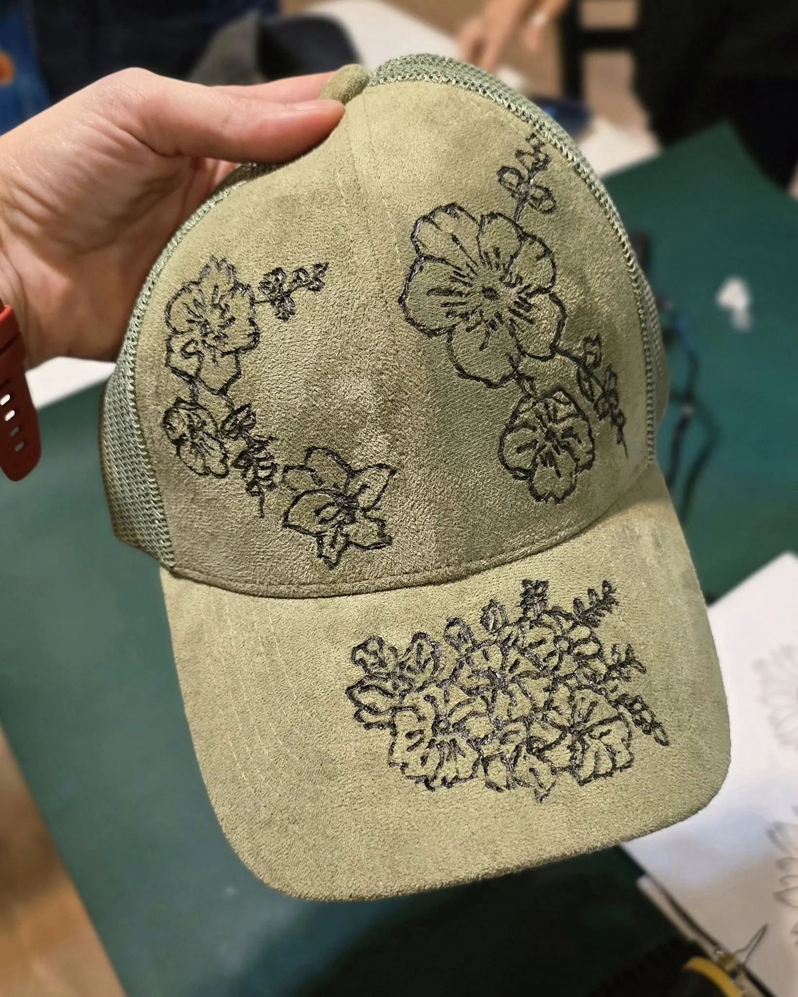 Close-up of a beige cap with hand-burnt black floral design on the front.