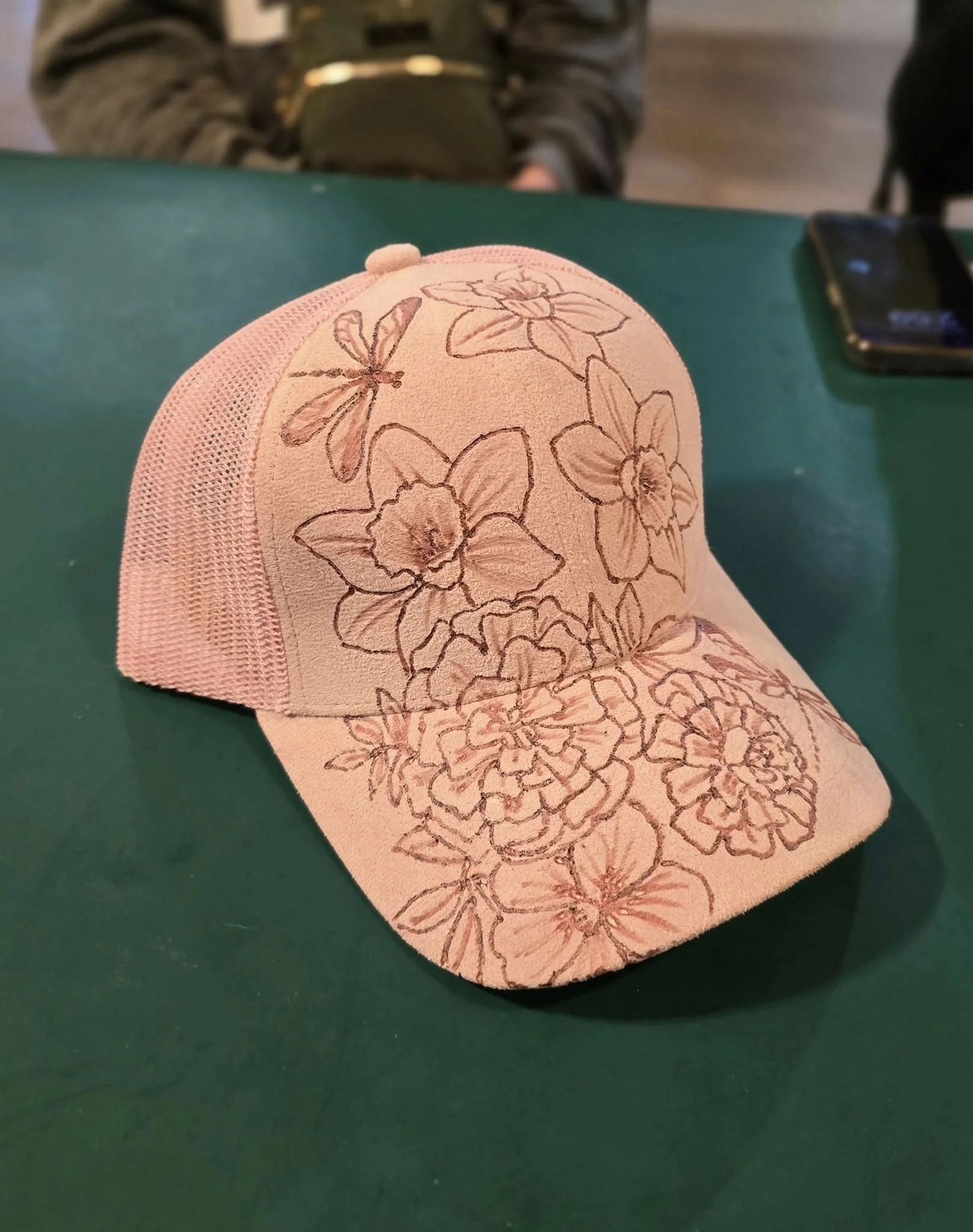 A pink baseball cap with floral hand-burnt design resting on a green surface, partially covering a black smartphone. In the background, a person in a striped sweater is visible, with their torso and arms out of focus.