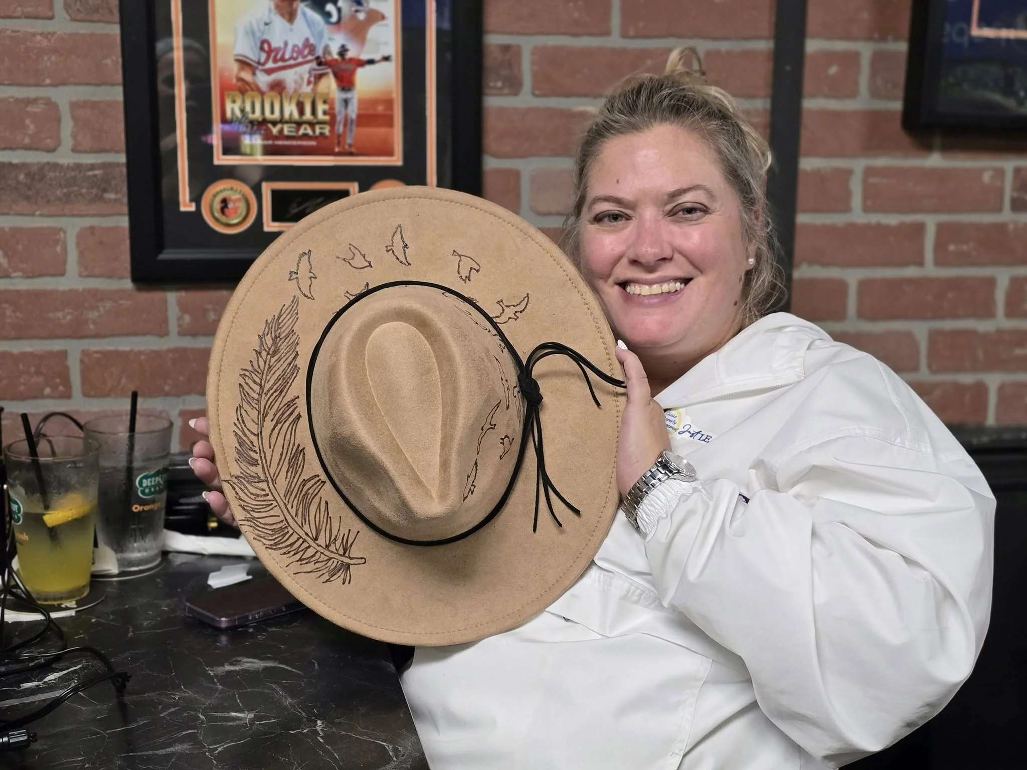 	
A woman smiling and holding a decorated cowboy hat in front of a brick wall and framed sports memorabilia. There are drinks on the table in front of her.