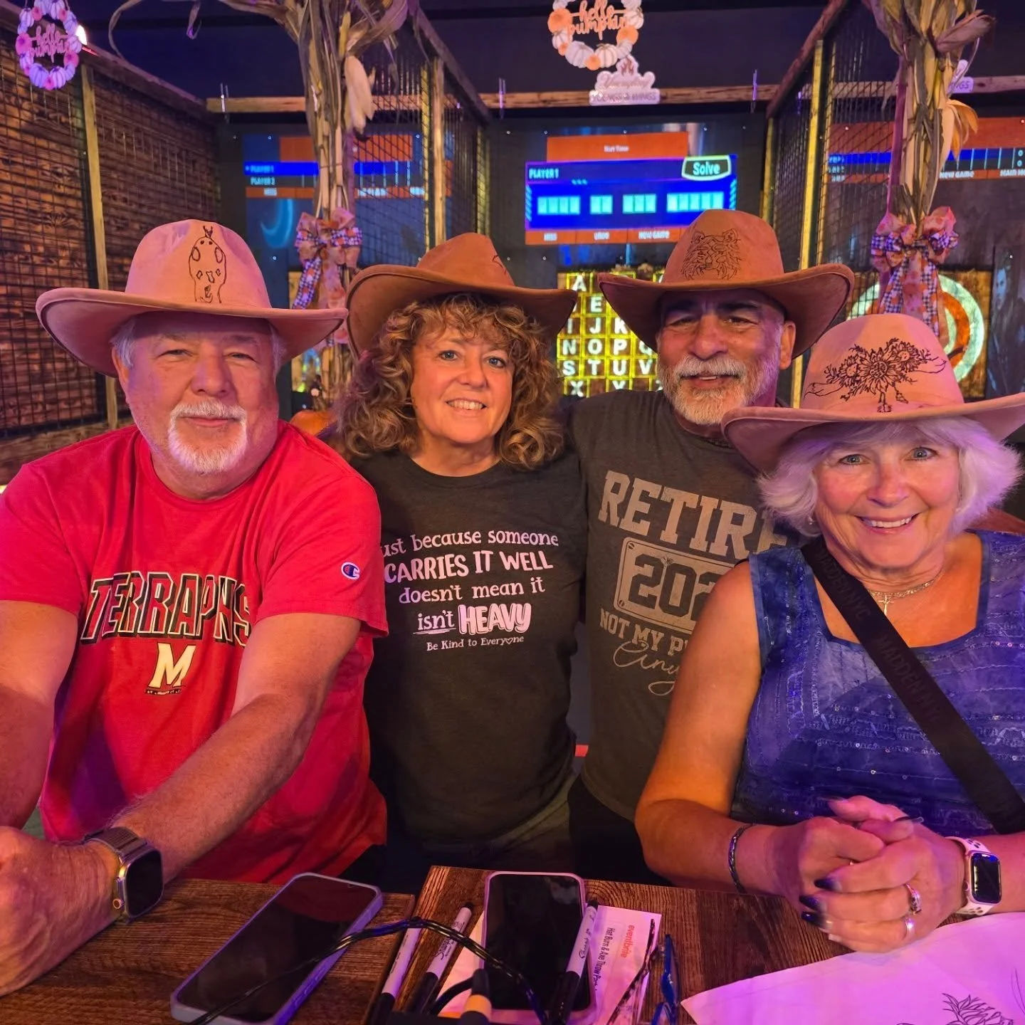 Four smiling adults wearing cowboy hats sitting at a table in a bar or restaurant with neon signs and electronic gaming screens in the background.