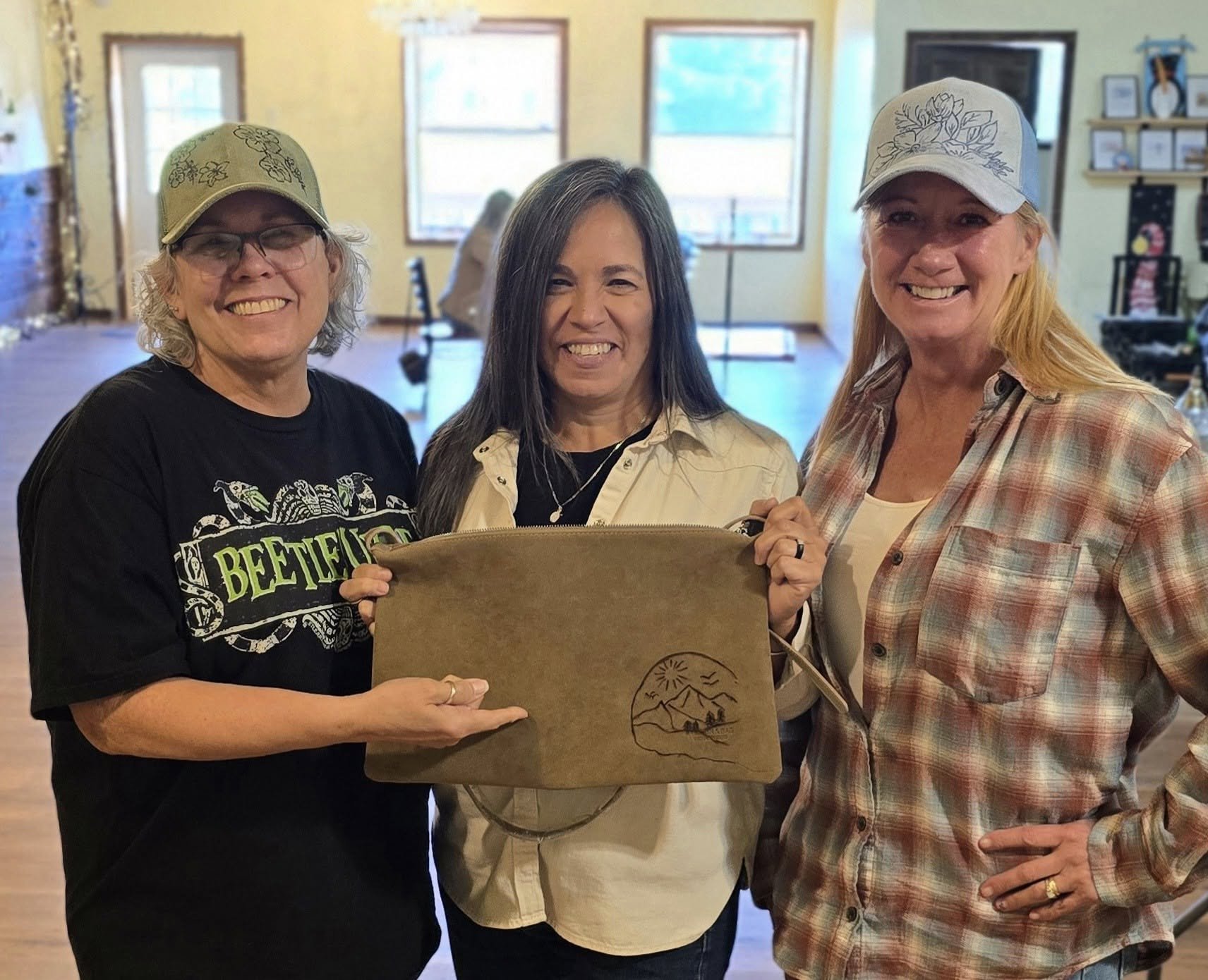 Three women smiling indoors, holding a brown purse with a mountain and sunset illustration.