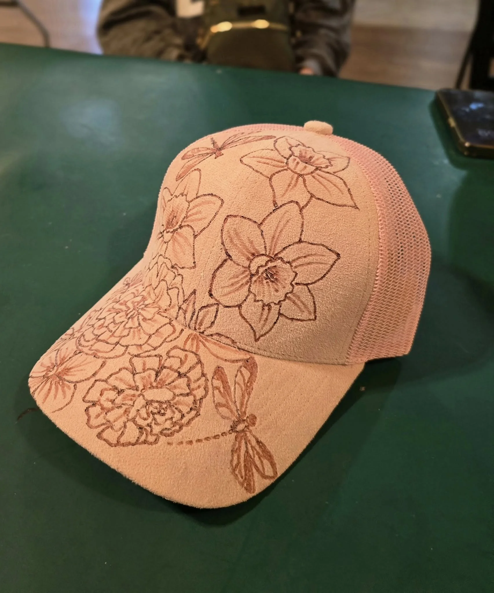 A close-up view of an orange-colored knit hat with hand-burnt floral design, resting on a green surface.