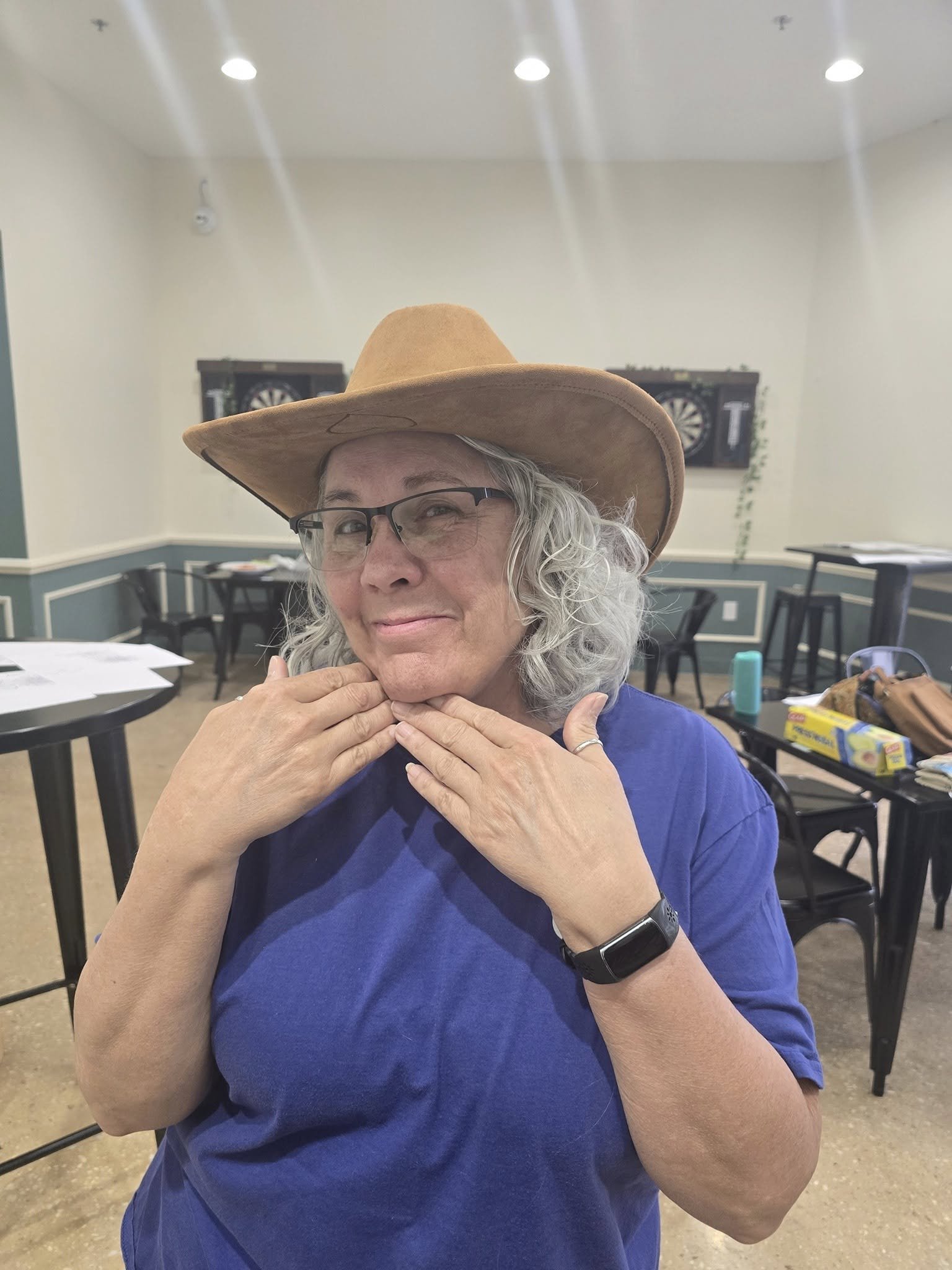 A woman with gray hair, glasses, and a tan wide-brimmed hat smiling and posing with her hands near her face in a room with dartboards on the wall and tables with various items.