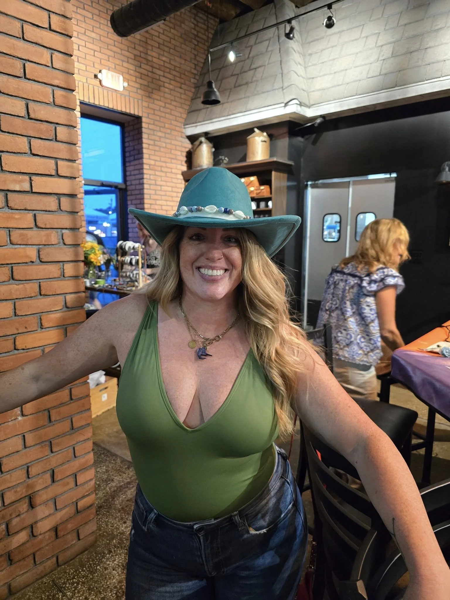 A smiling woman with long wavy blonde hair wearing a large teal hat decorated with shells and beads, a green sleeveless top, and blue jeans, seated at a table inside a restaurant with brick and concrete walls, and other people in the background.