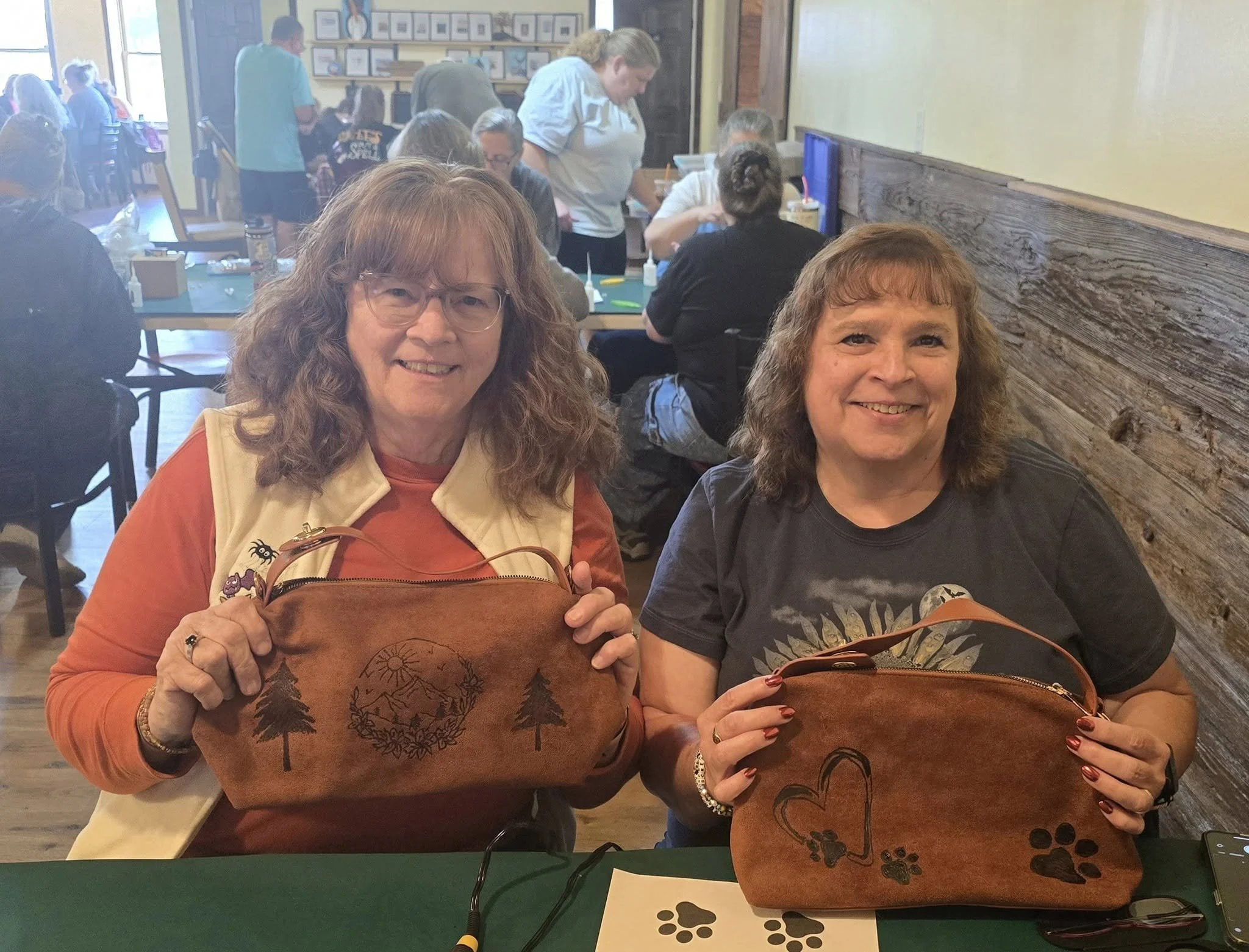 Two women smiling and holding up brown leather bags with mountain and tree designs in a cozy restaurant or cafe setting, with people in the background.
