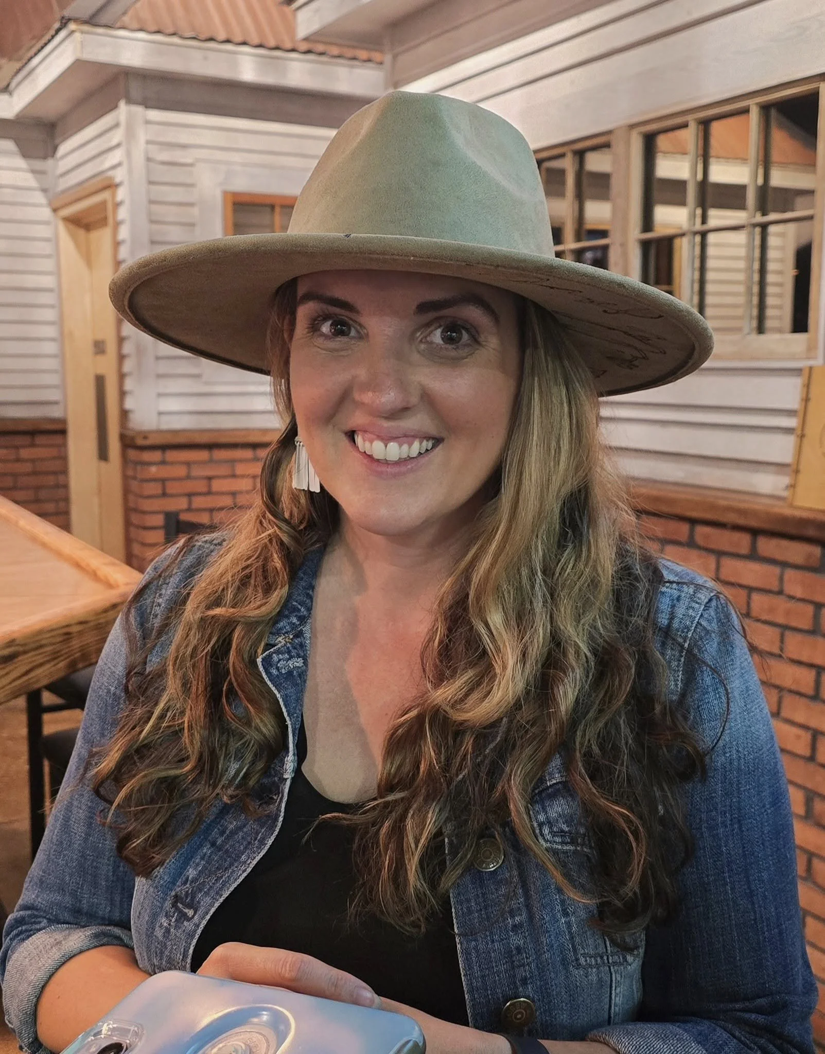 A woman with long wavy hair, smiling, wearing a wide-brimmed hat and denim jacket, sitting at a table indoors with wooden and brick architectural elements in the background.