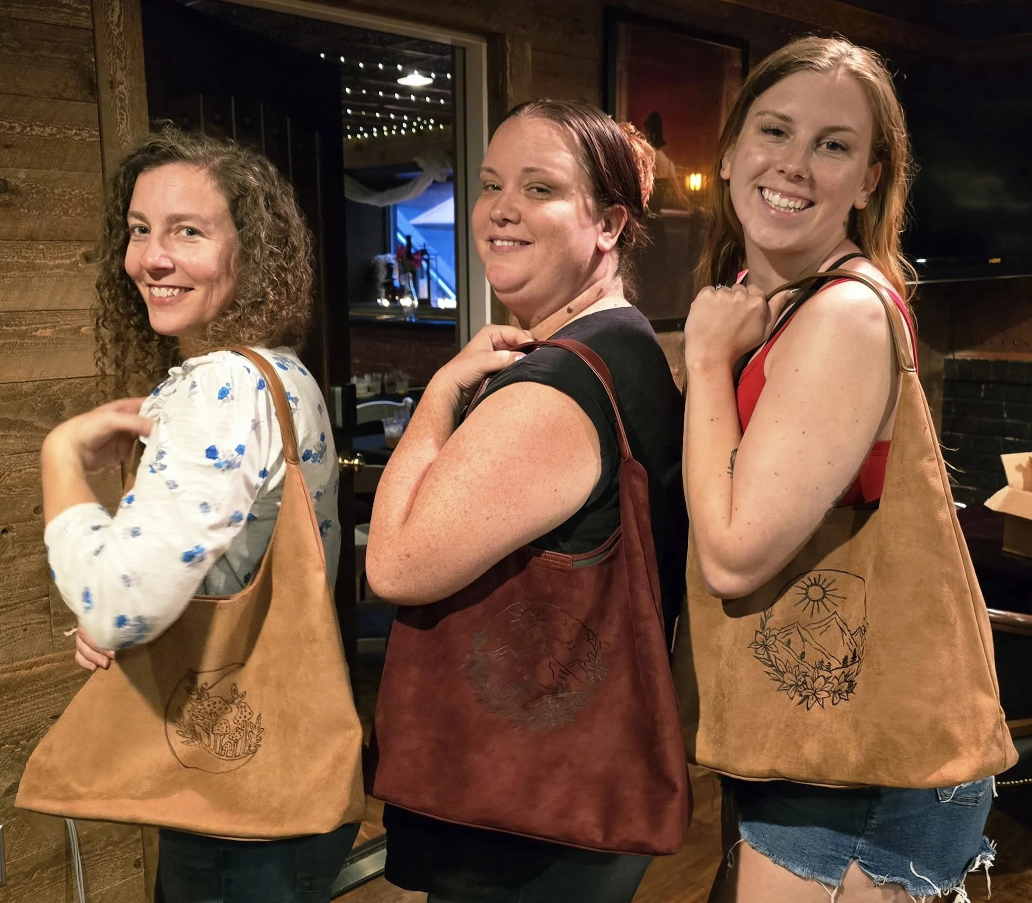 Three women standing in a row, smiling and looking at the camera, inside a wooden interior space, each carrying a brown bag with a burnt design, with a large window showing outdoor string lights.