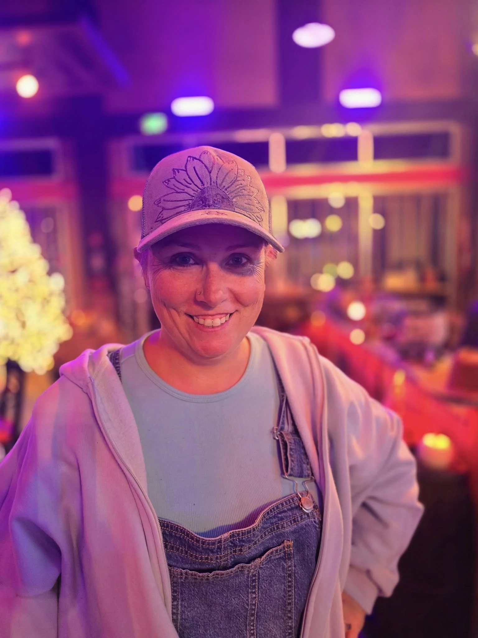 A woman smiling in a colorful, neon-lit indoor setting, wearing a baseball cap with a floral design, a light-colored hoodie, and denim overalls.
