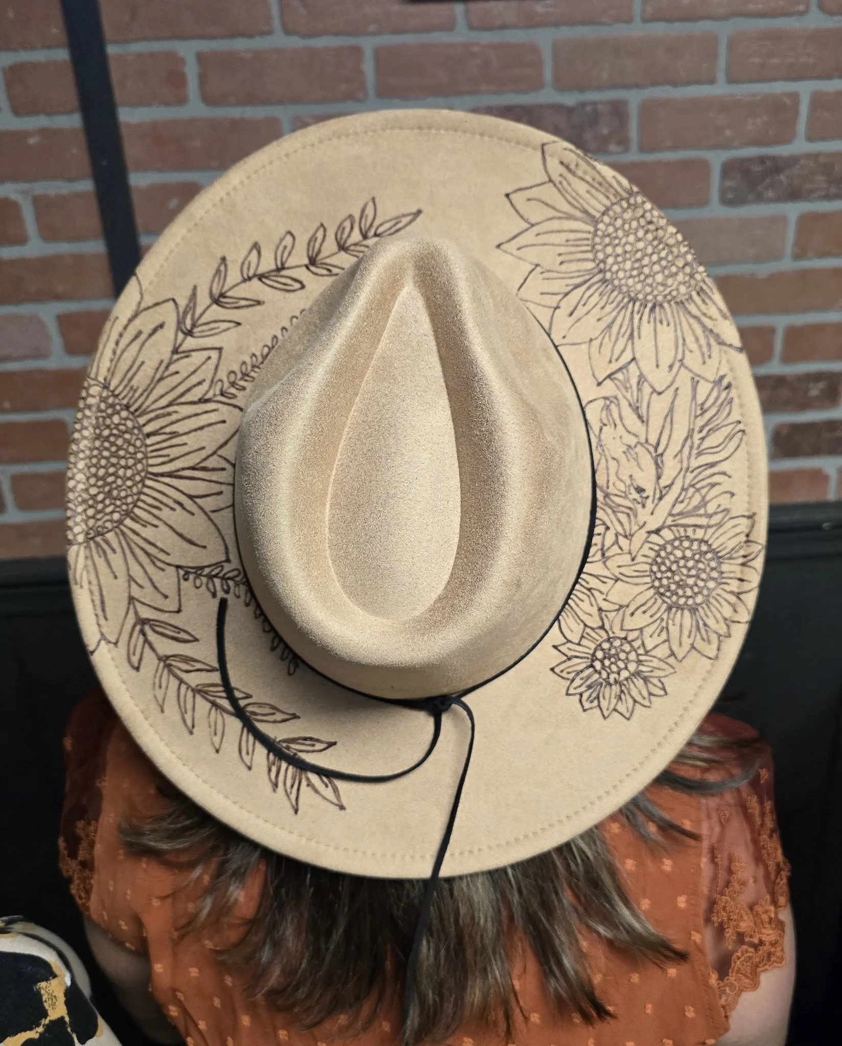 A tan western-style hat with hand-burnt floral and leaf designs, featuring resting on a person's head against a brick wall background.