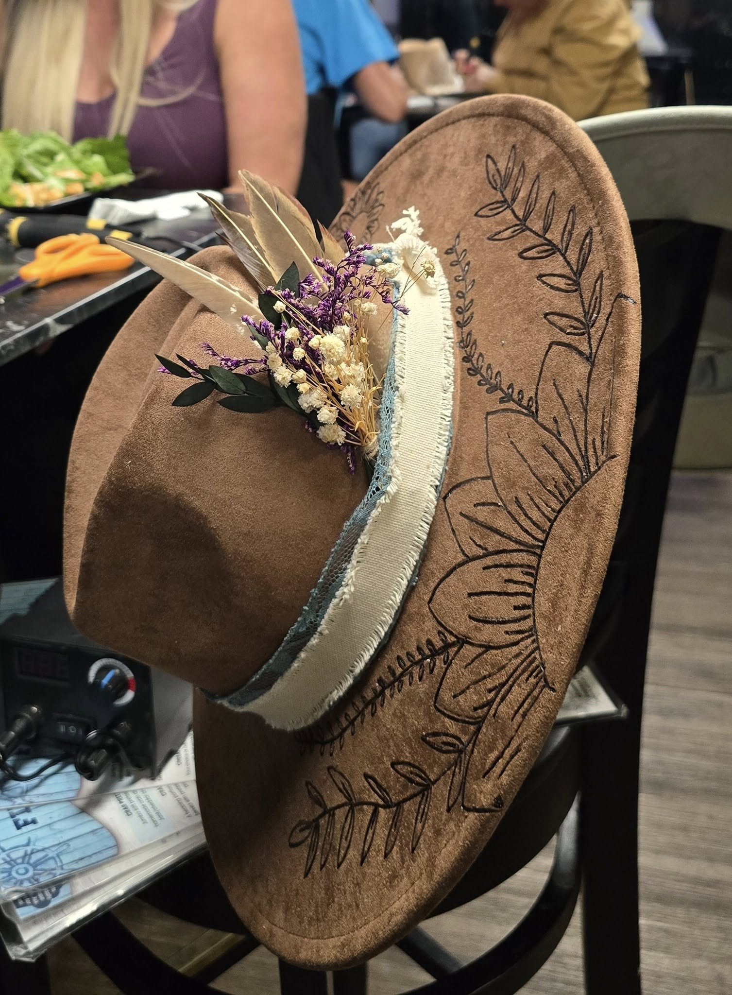 A close-up of a brown cowboy hat with black burnt leaf designs, placed on a chair at a table in a restaurant. The hat has decorative feathers and a small bouquet of white and purple flowers attached to the band.